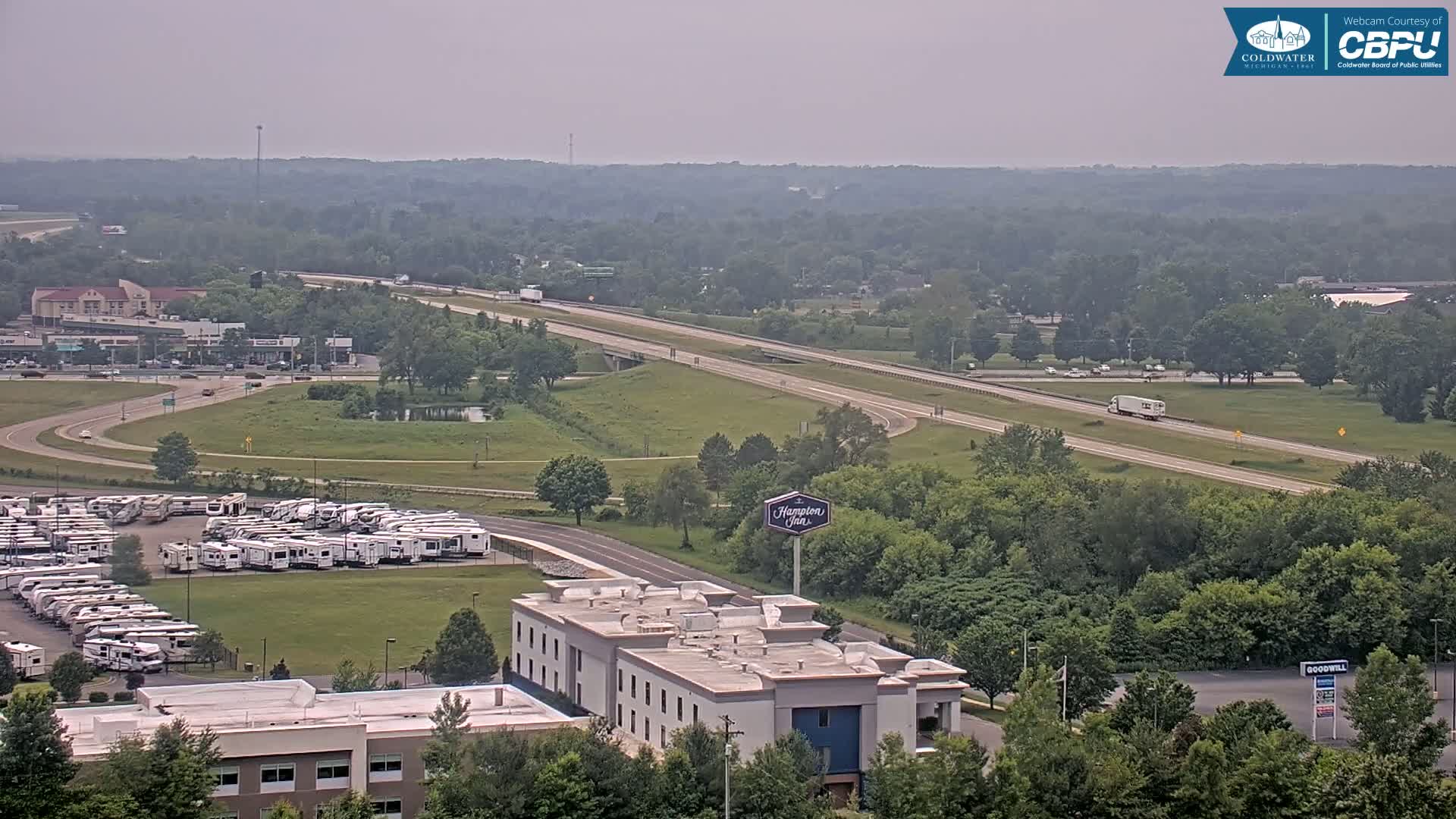 A hazy view of a highway, a large RV parking lot, a Hampton Inn, and surrounding green landscape.