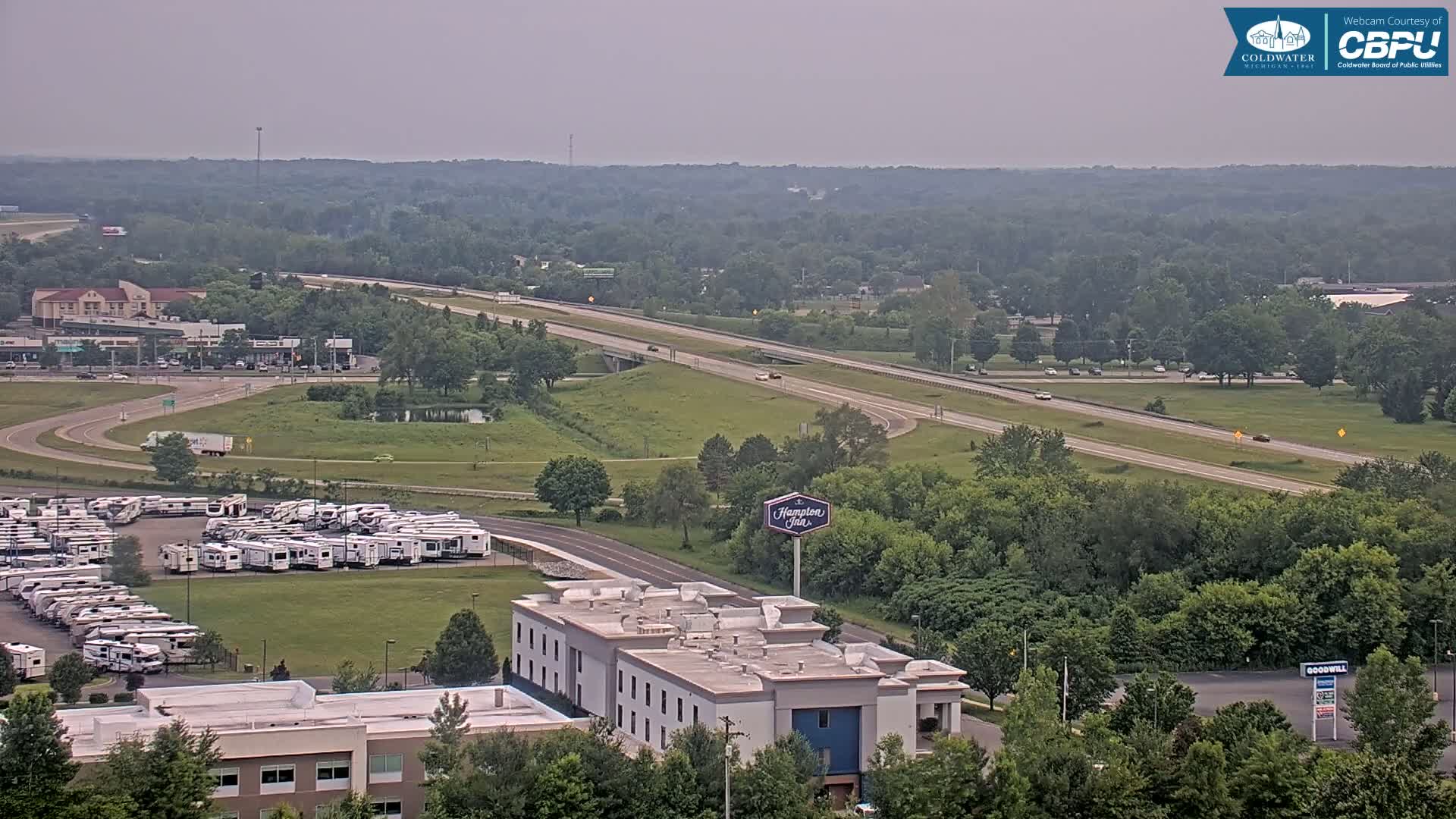 A hazy view of a highway, a large RV parking lot, a Hampton Inn, and surrounding green landscape.