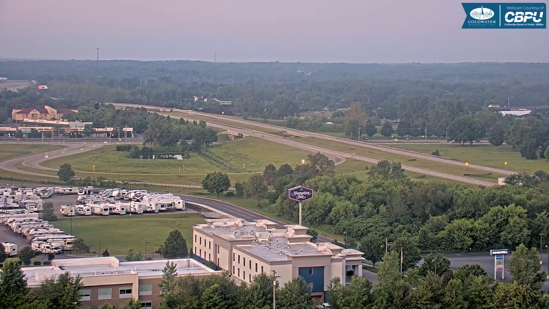 An elevated view of a highway, a large RV park, a Hampton Inn, and a surrounding landscape of green trees and fields under a hazy sky.