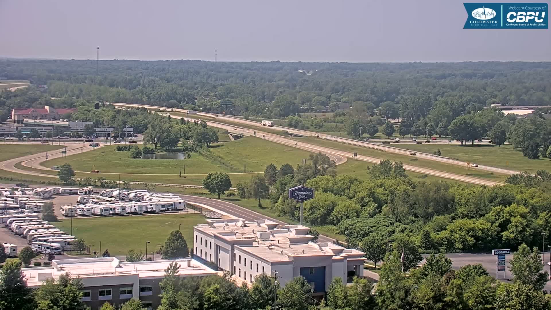 A high-angle, long shot on a sunny day shows a highway cutting through a green landscape with a parking lot full of RVs, and a Hampton Inn visible near the bottom.