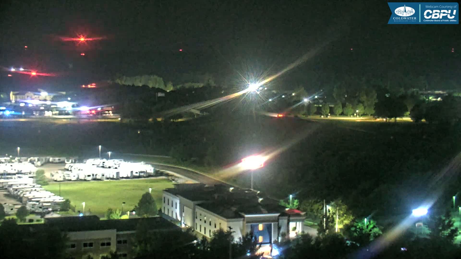 A nighttime aerial view shows a motel, a large RV park, and a roadway, all surrounded by dark green trees under a clear sky.