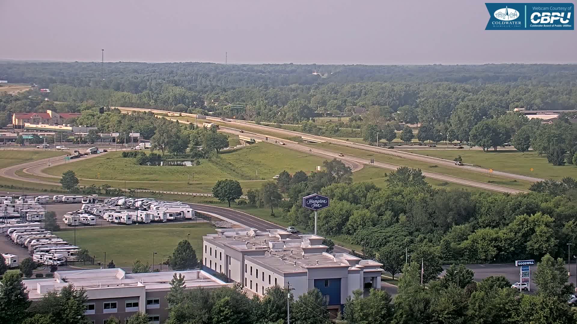 A high-angle, long shot shows a highway, a large parking lot filled with RVs, a Hampton Inn, and surrounding green landscape on a hazy day.