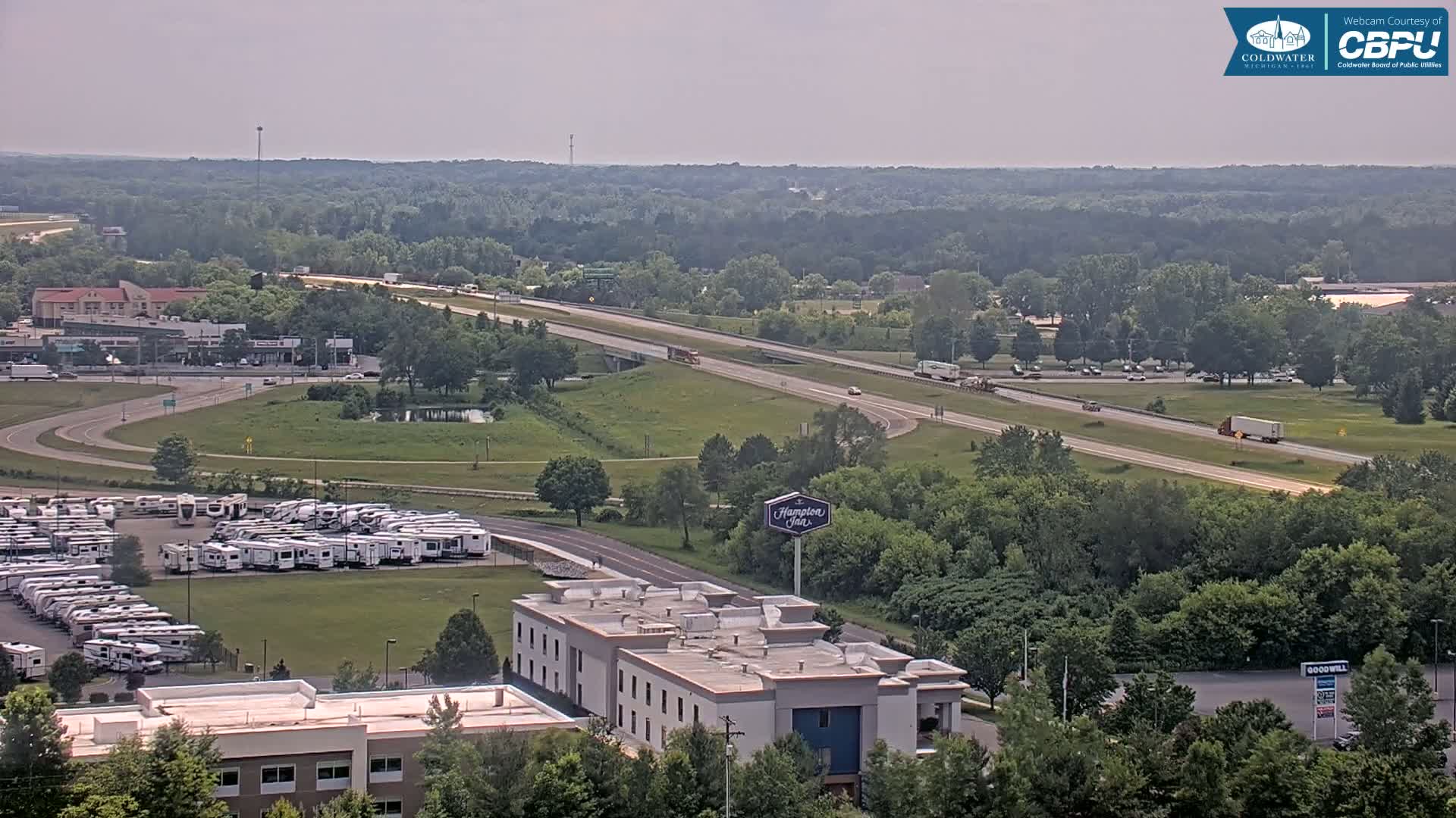 A mostly sunny view of a highway, a large RV park, a Hampton Inn, and surrounding greenery.