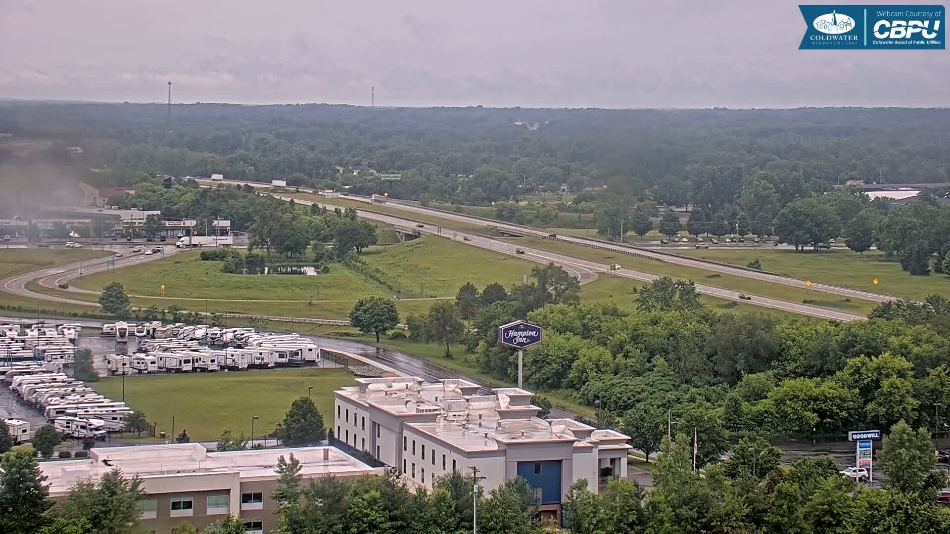 An aerial view shows a highway with sparse traffic, a large RV park, a Hampton Inn, and surrounding green space under an overcast sky.