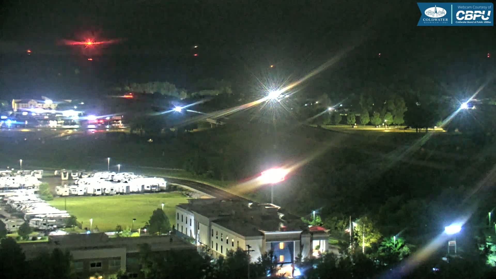 A nighttime aerial view shows a large RV park, a hotel, and surrounding landscape illuminated by lights, with several distant red lights possibly from aircraft or emergency vehicles in the sky.