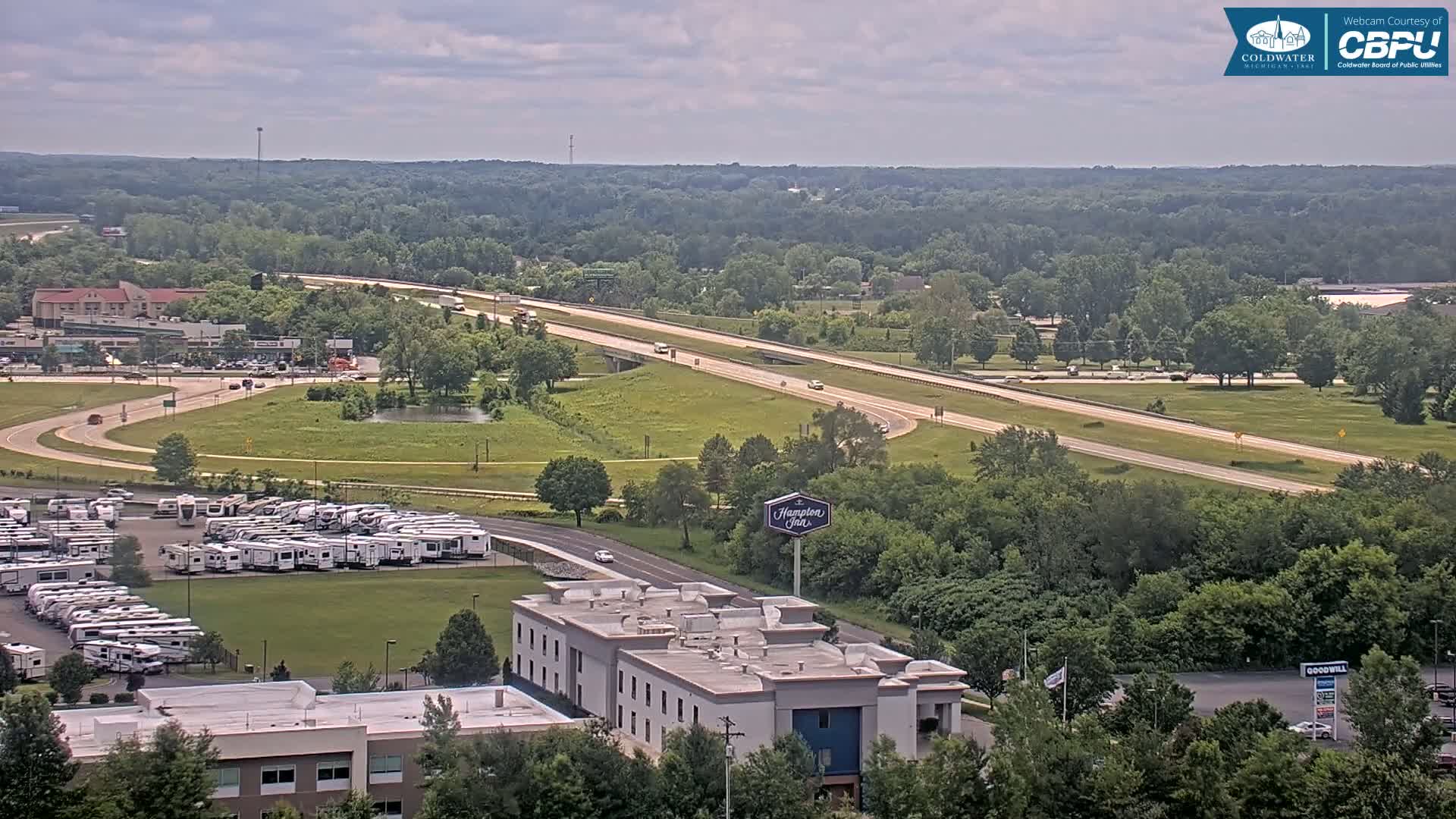 A partly cloudy day overlooking a highway, a large RV park, and a Hampton Inn, surrounded by green trees and grassy areas.