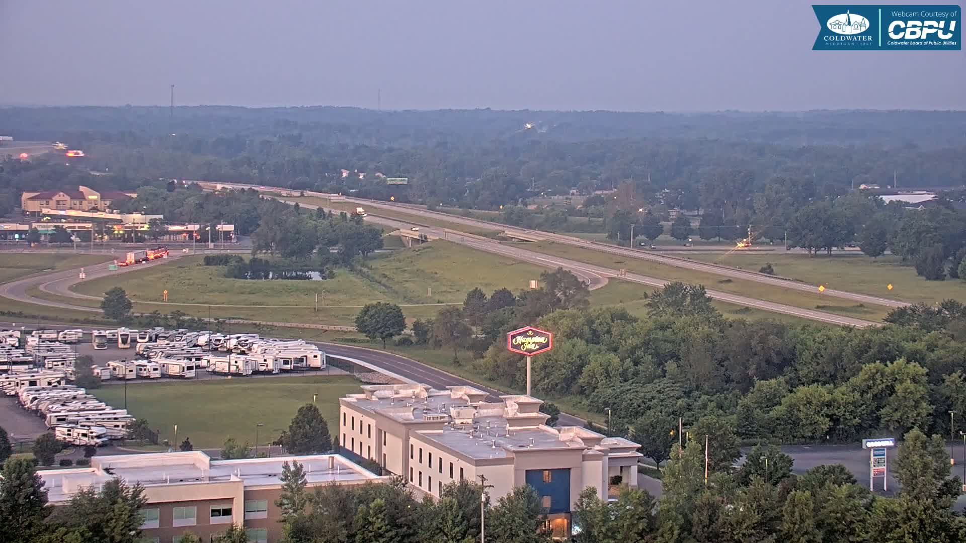 An aerial view of a highway, a large RV park, a Hampton Inn hotel, and surrounding green spaces under a dusky sky.