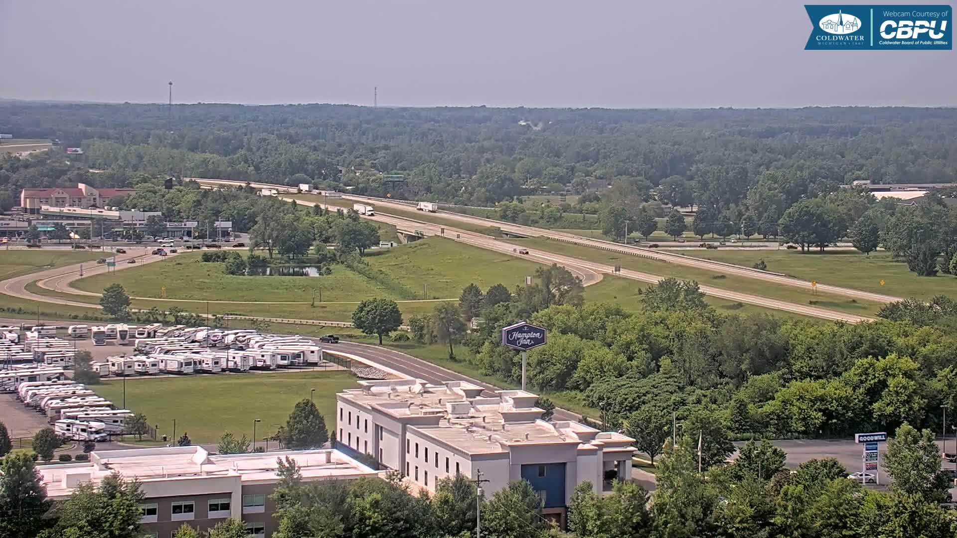 A highway stretches through a green landscape under a clear, sunny sky, with a large RV park and a hotel visible near the road.