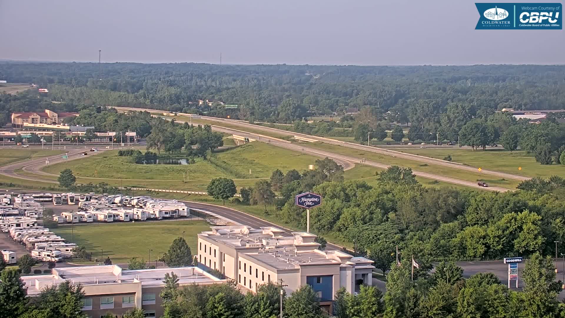 A high-angle, long shot shows a highway, a hotel, and a large RV park nestled within a landscape of green trees and fields under a hazy sky.