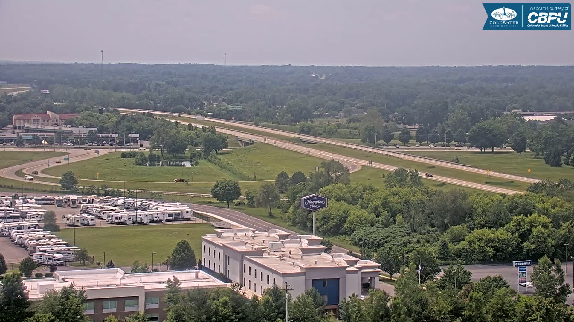 A highway stretches through a green landscape, passing near a large RV park and a hotel, under a hazy, sunny sky.