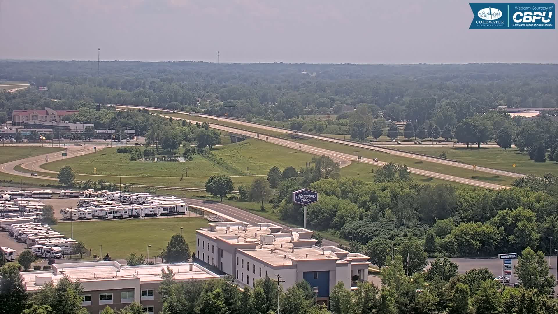 A highway stretches through a landscape of green trees and grassy areas, with a Hampton Inn and a large RV parking lot visible nearby, under sunny skies.