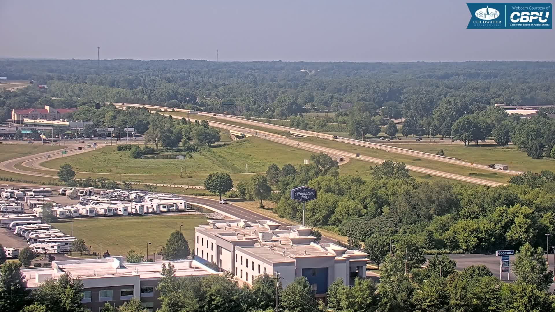 A highway stretches through a green landscape with a Hampton Inn and a large RV parking lot visible, under sunny skies.