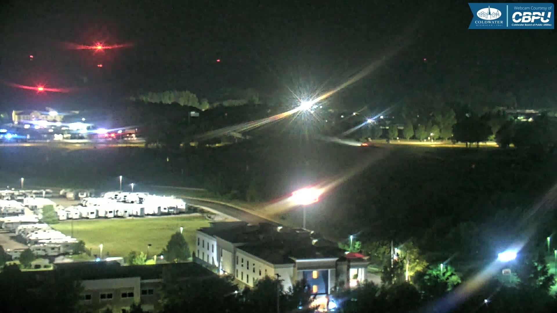 A nighttime aerial view shows a parking lot filled with RVs, a hotel, and a road with vehicle lights, under a clear, dark sky with distant red lights.