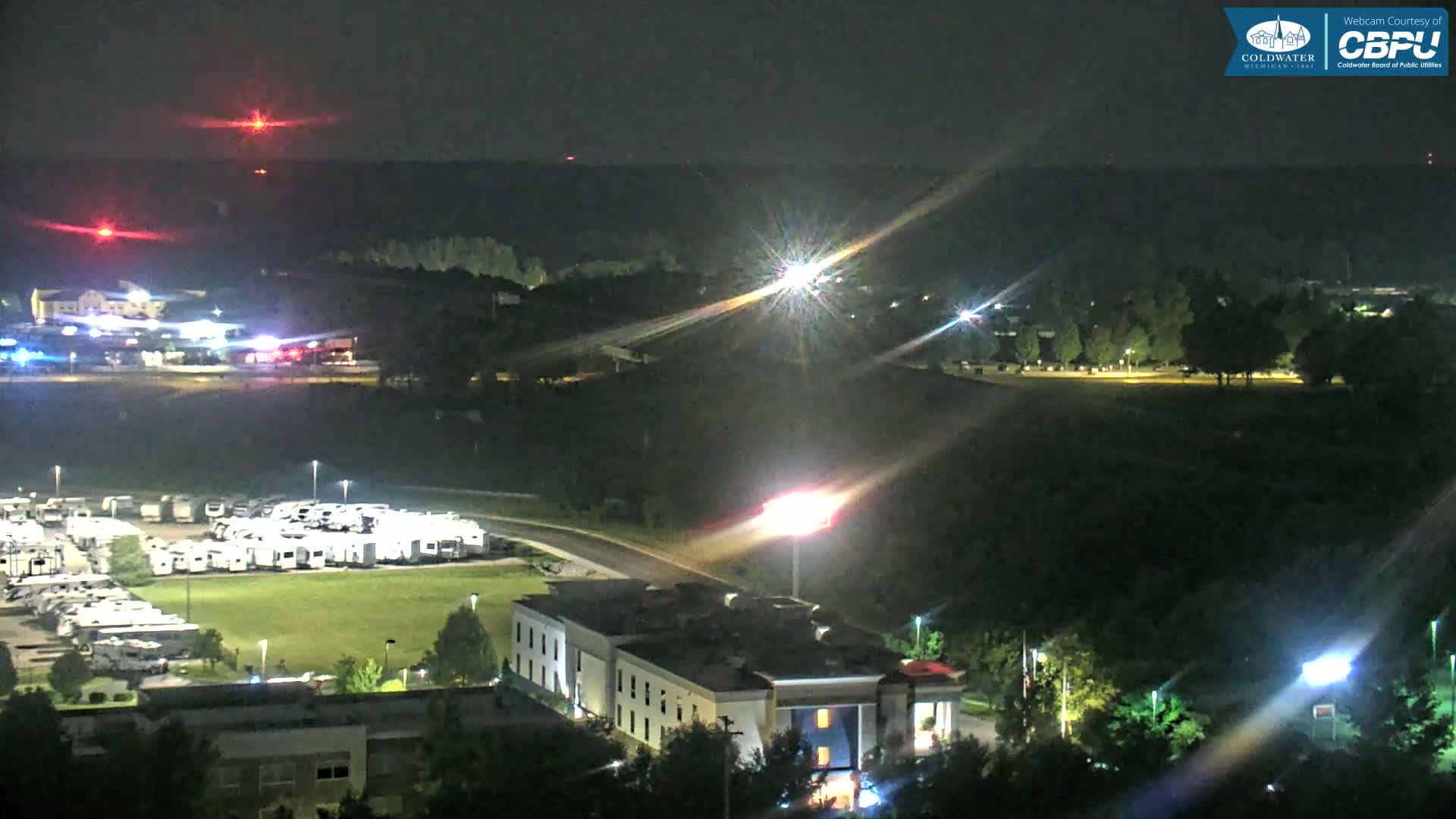 A nighttime aerial view shows a parking lot filled with RVs, a hotel, and a road with vehicle lights, under a clear, dark sky with distant red lights.