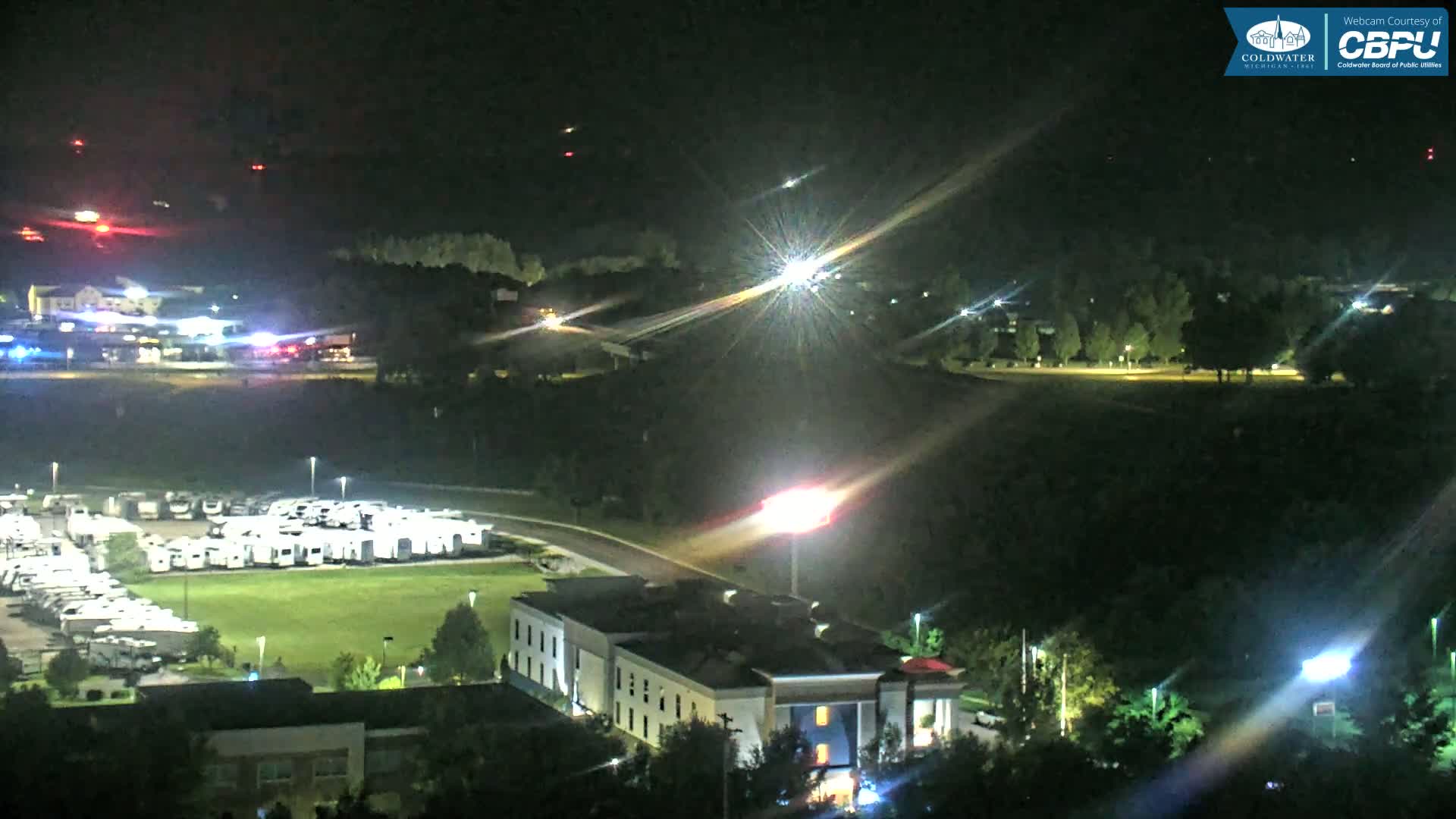 A nighttime aerial view shows a parking lot filled with RVs, a hotel, and a road with vehicle lights, under a clear, dark sky with distant red lights.