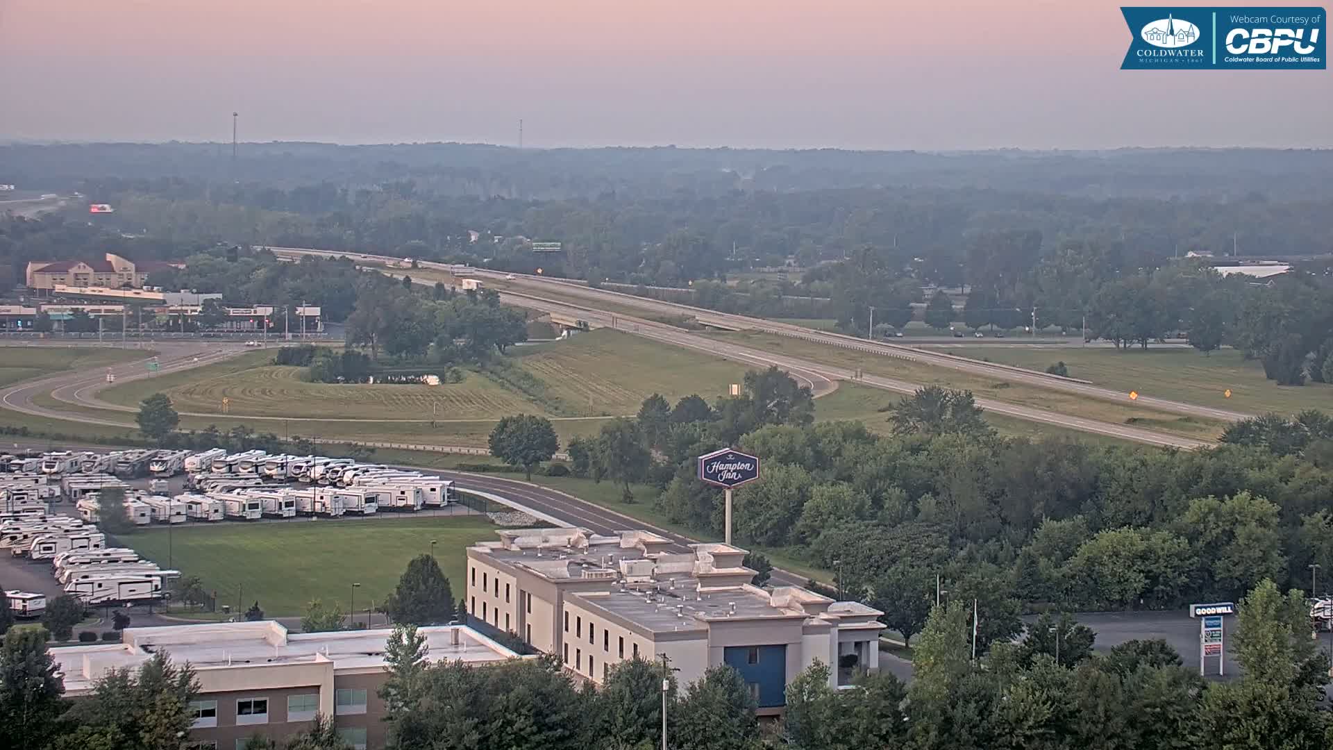 A nighttime aerial view shows a parking lot filled with RVs, a hotel, and a road with vehicle lights, under a clear, dark sky with distant red lights.