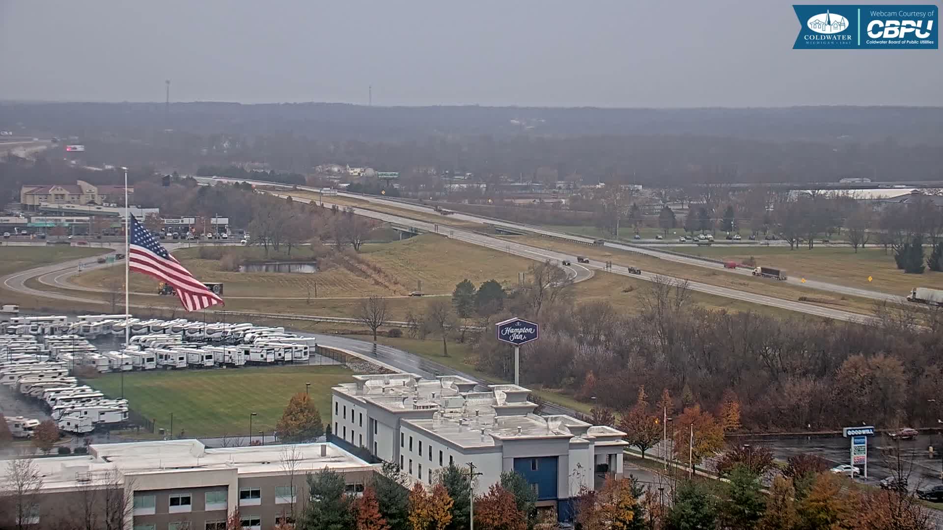 A nighttime aerial view shows a parking lot filled with RVs, a hotel, and a road with vehicle lights, under a clear, dark sky with distant red lights.
