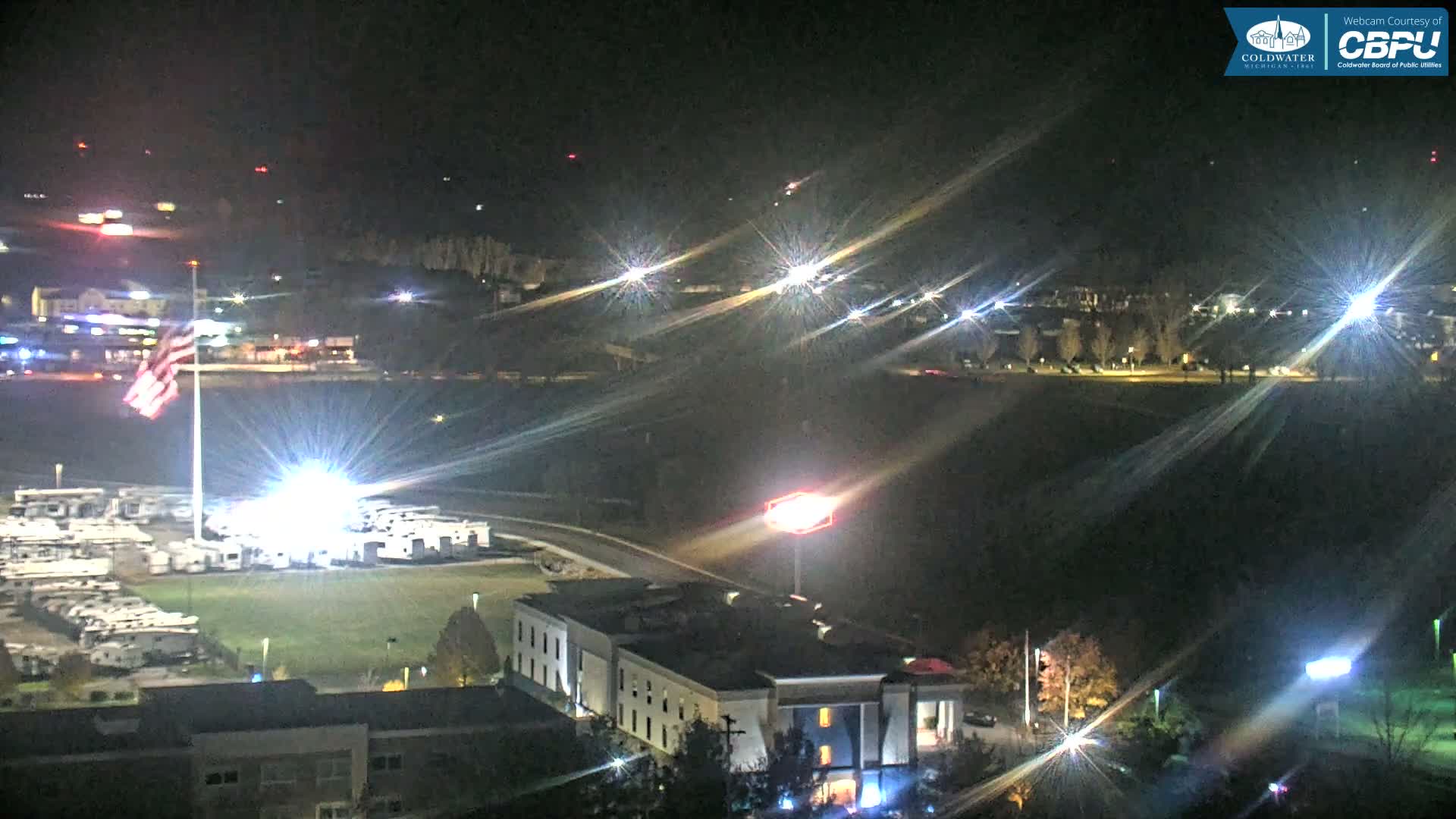 An illuminated American flag waves over a parking lot filled with RVs and various buildings, with distant city lights and a highway visible under a clear night sky.