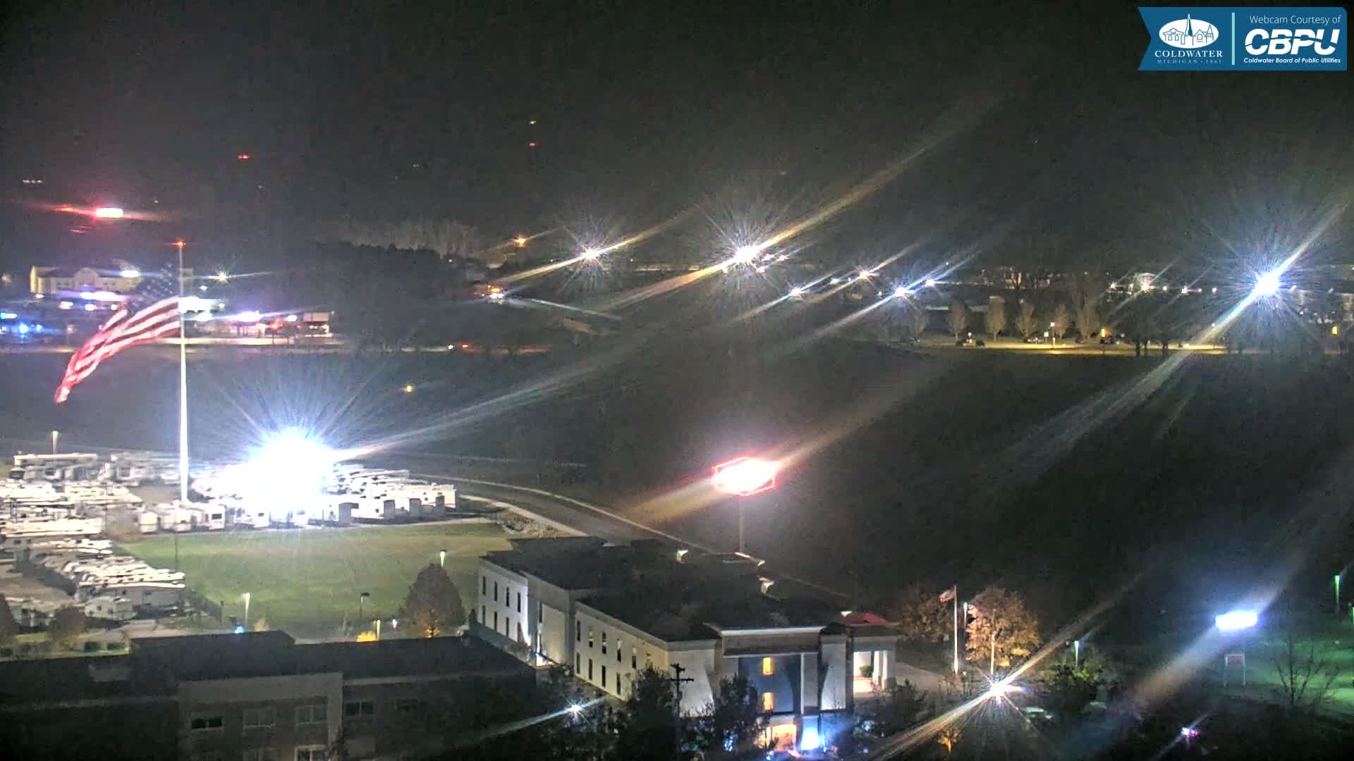 A clear nighttime scene captures a large American flag illuminated by a brilliant spotlight, overlooking a lot of RVs and several buildings, with numerous sparkling distant lights from roads and structures visible in the background.