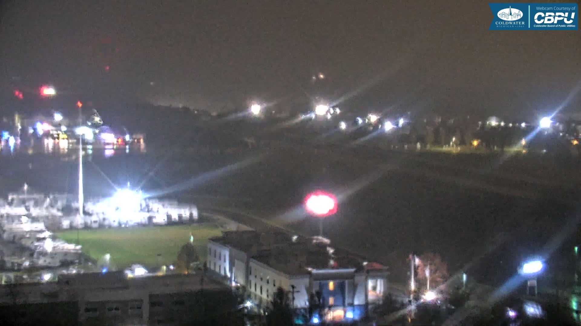 An illuminated townscape at night features a body of water reflecting numerous colorful lights, brightly lit buildings, and a prominent glowing red sign, all under hazy, overcast conditions.