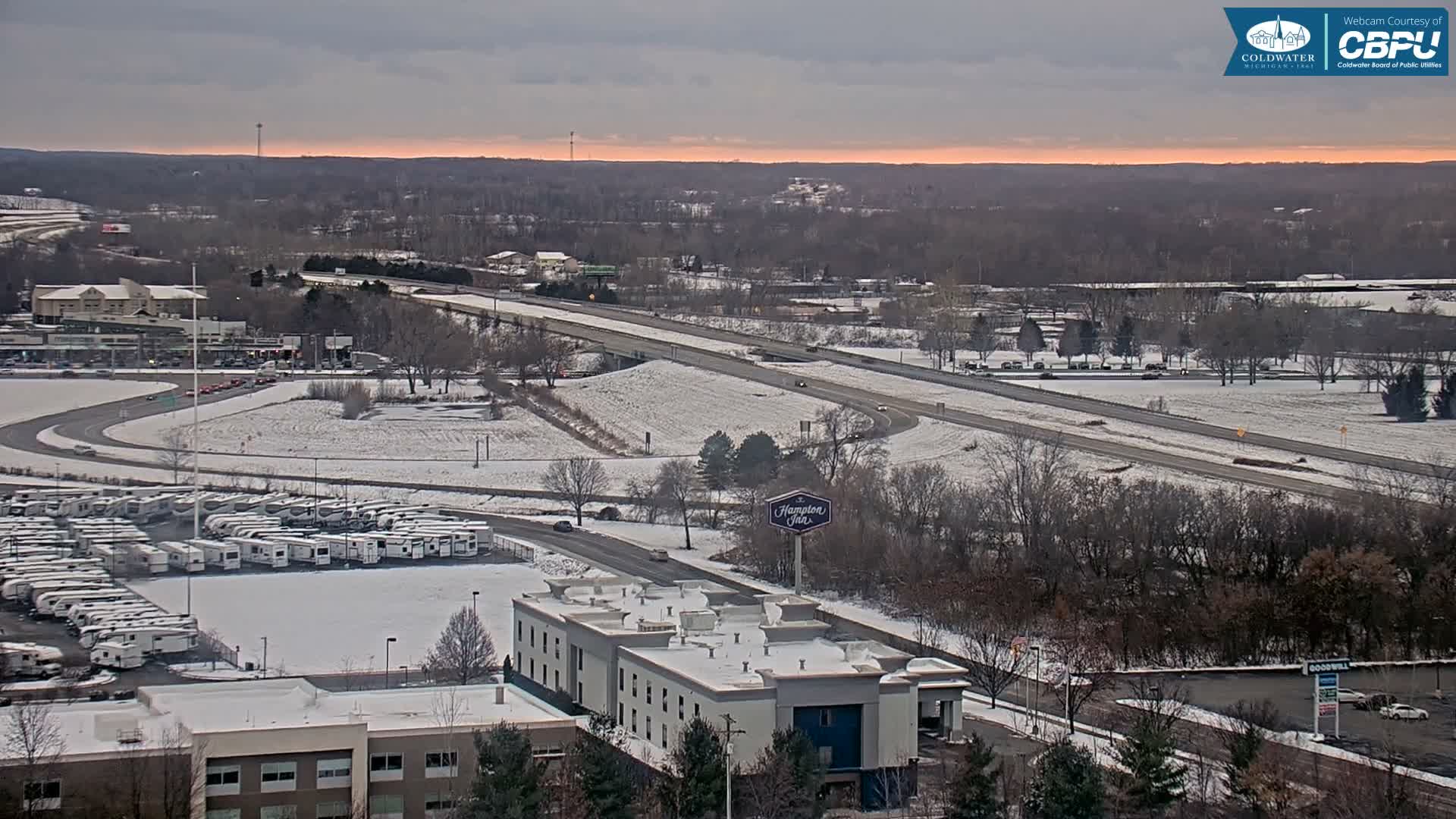 A wintry, overcast scene reveals a snow-dusted urban landscape featuring a multi-lane highway, commercial buildings, and a large RV parking lot, all under a gray sky with a faint orange glow on the distant horizon.