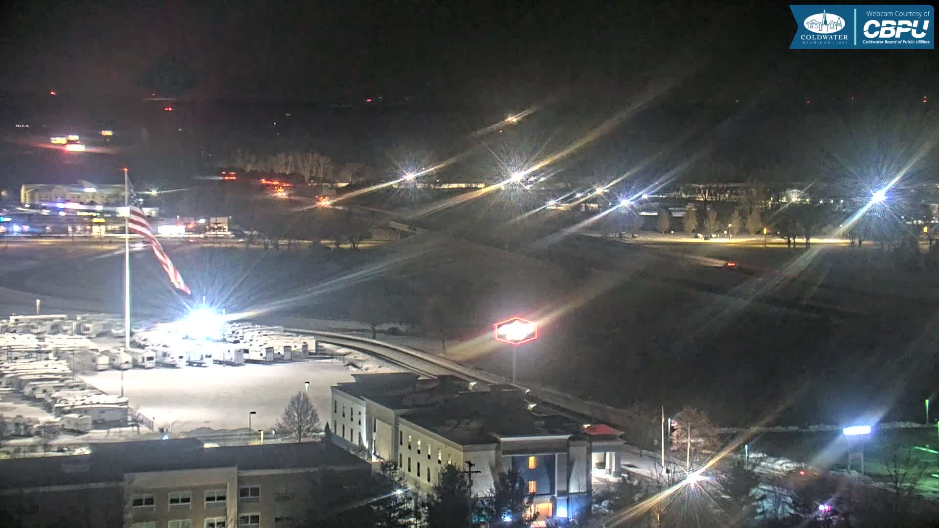 On a clear, snowy winter night, an illuminated American flag stands over a snow-covered lot of parked RVs, with buildings and distant lights visible across the landscape.
