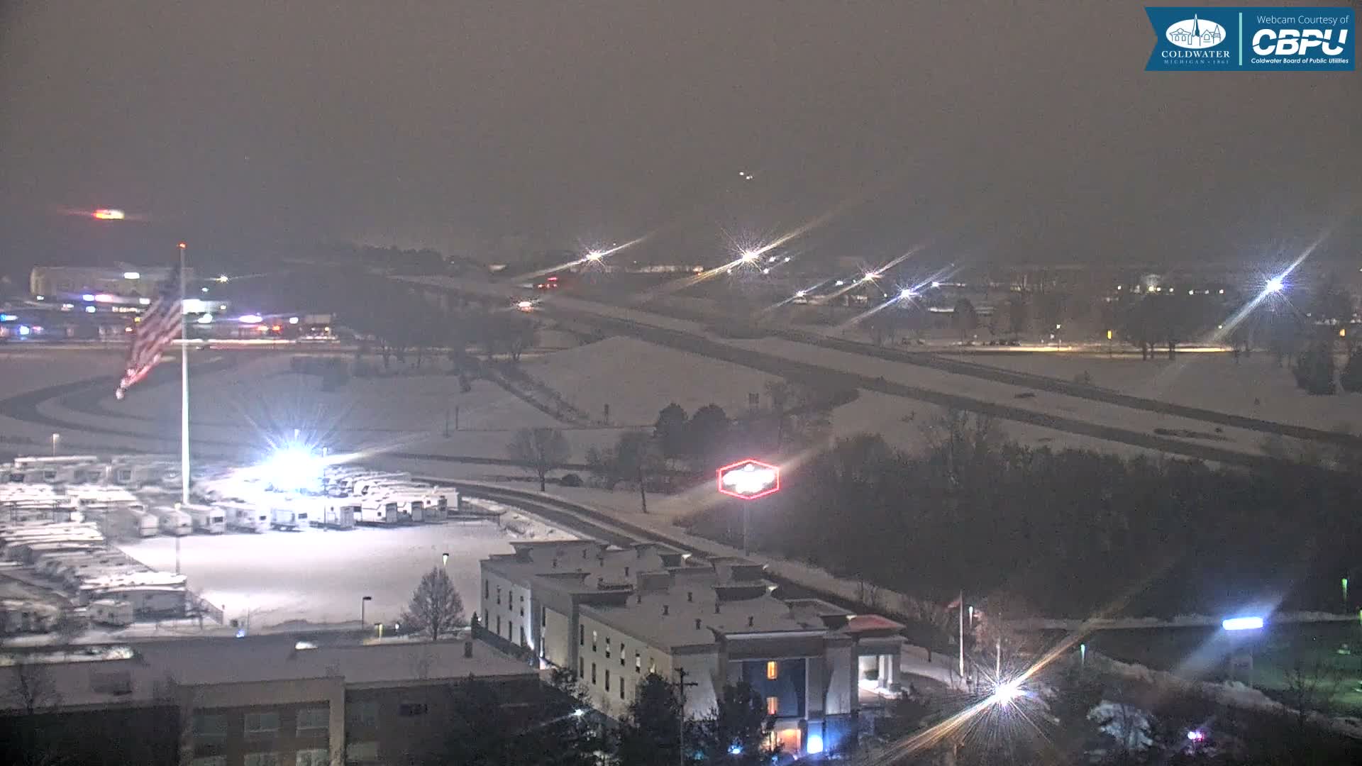 An overcast and snowy winter night scene displays an illuminated American flag, a large parking lot full of RVs, several snow-covered buildings, and a distant highway dotted with vehicle lights.
