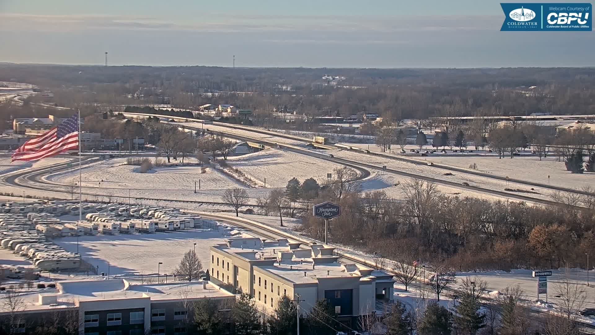 An overcast and snowy winter night scene displays an illuminated American flag, a large parking lot full of RVs, several snow-covered buildings, and a distant highway dotted with vehicle lights.