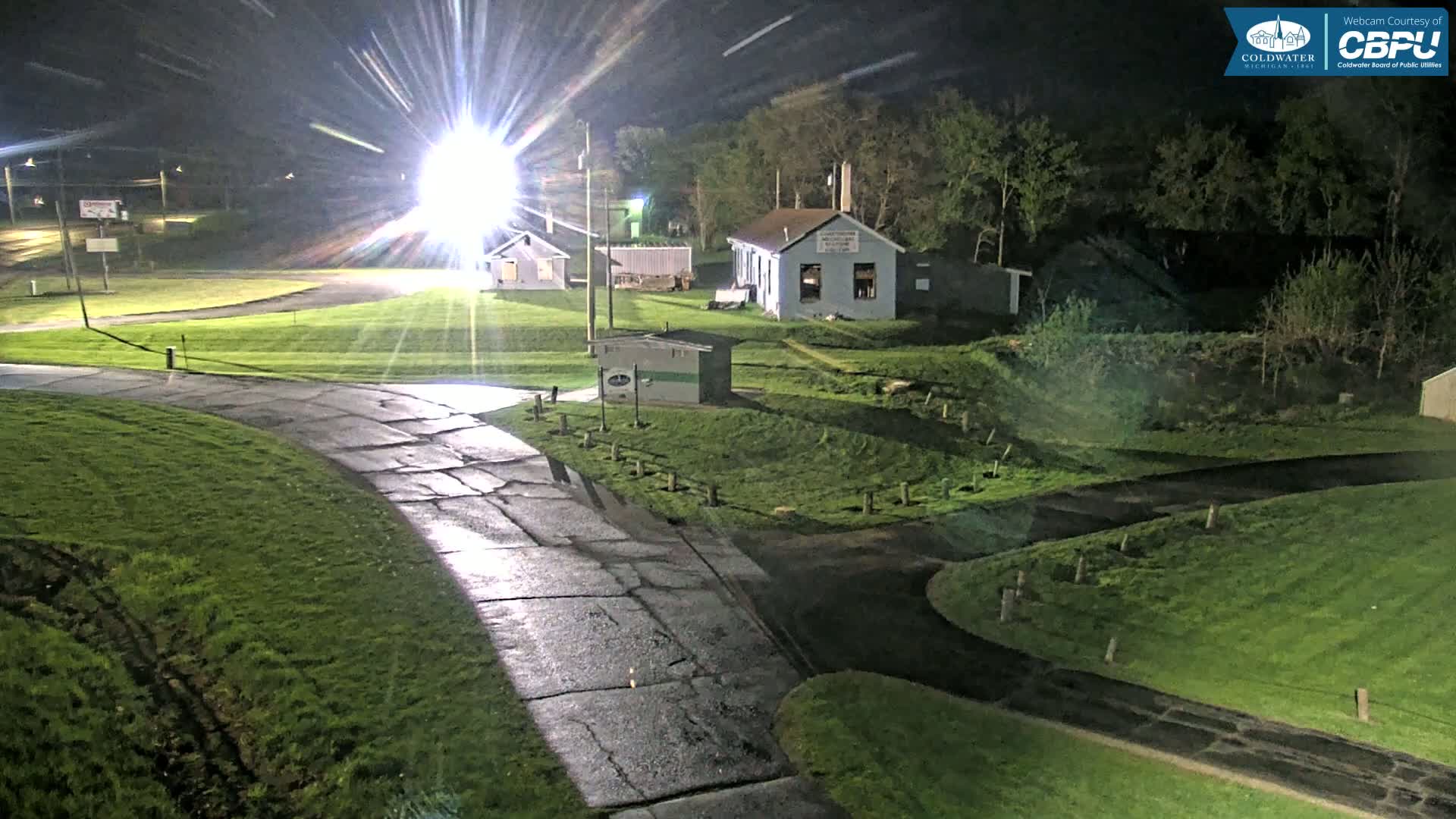 A nighttime view of a grassy area with paved roads and small buildings, lit by bright lights under a mostly clear sky.