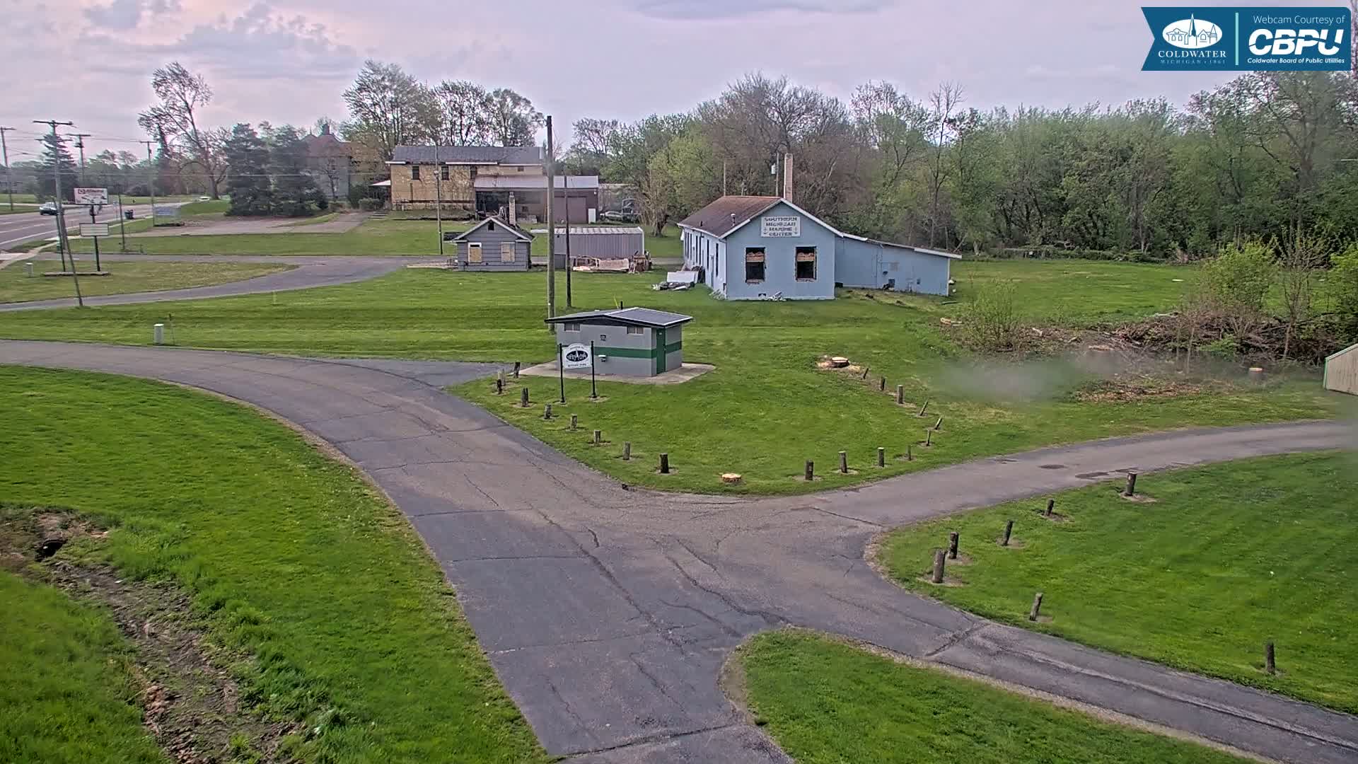 A paved road intersects with other paths in a grassy area, near several buildings under a cloudy sky.