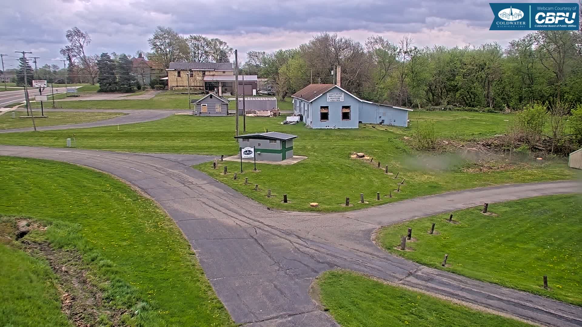 A paved road curves through a grassy area with several small buildings and trees under a partly cloudy sky.