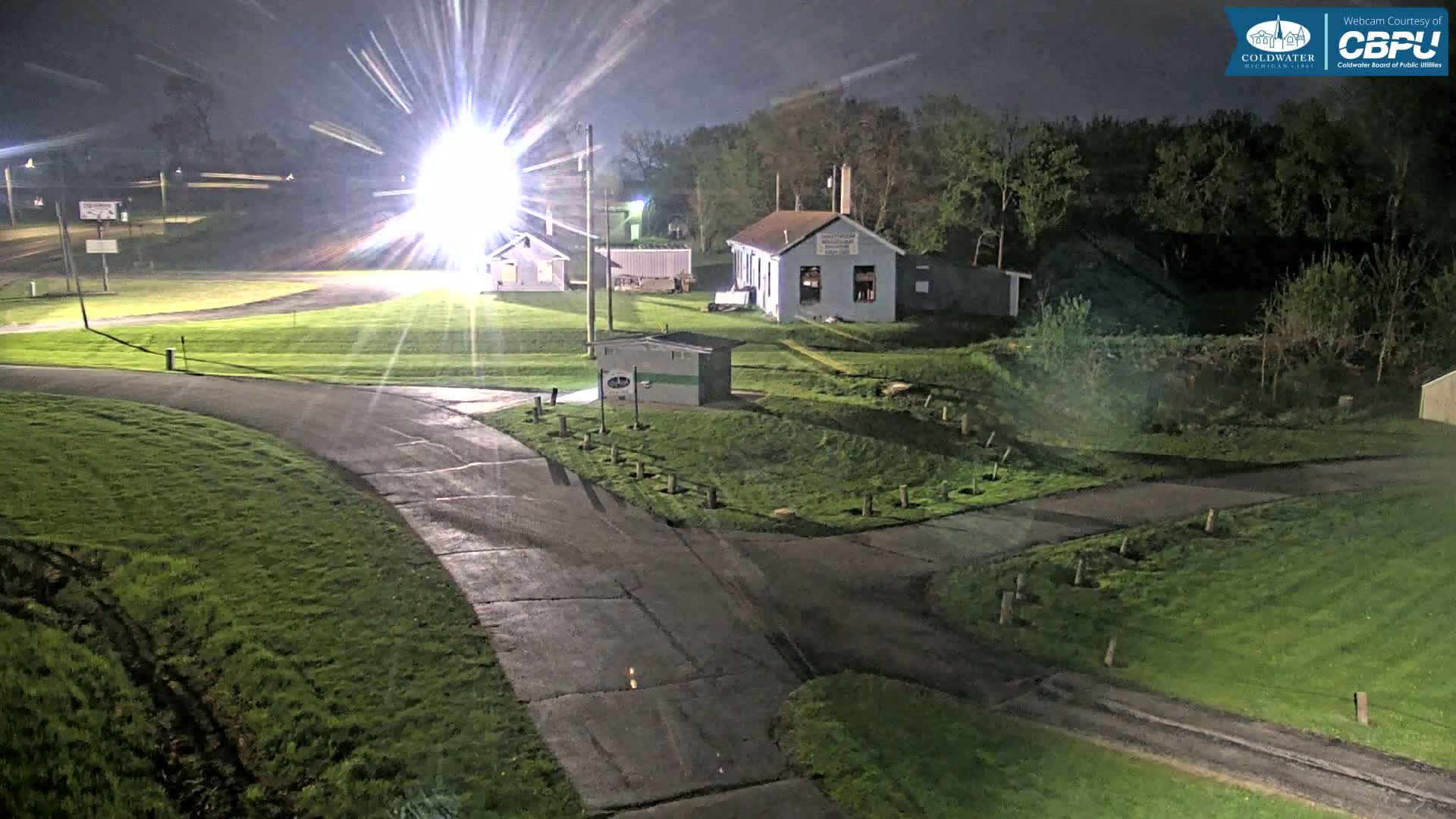 At night, under bright lights, paved paths wind through grassy areas past small buildings and trees.