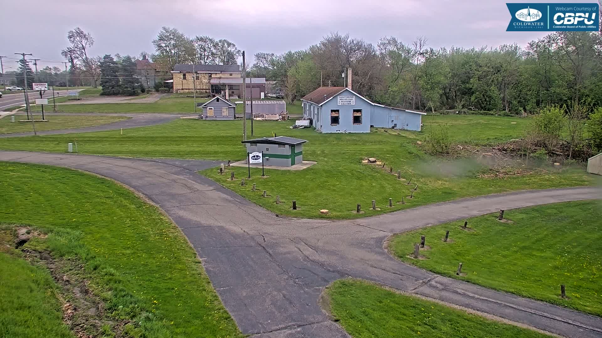 A paved, curving road intersects a grassy area with several small buildings under an overcast sky.