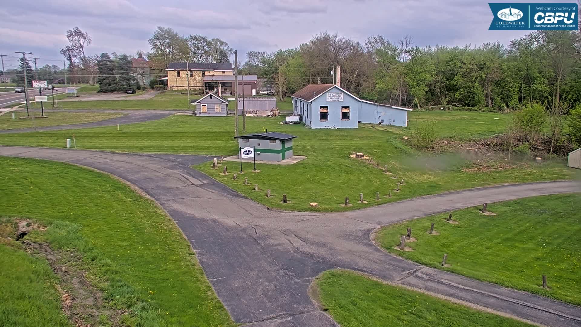 A paved roadway curves through a grassy area with several small buildings and trees under an overcast sky.