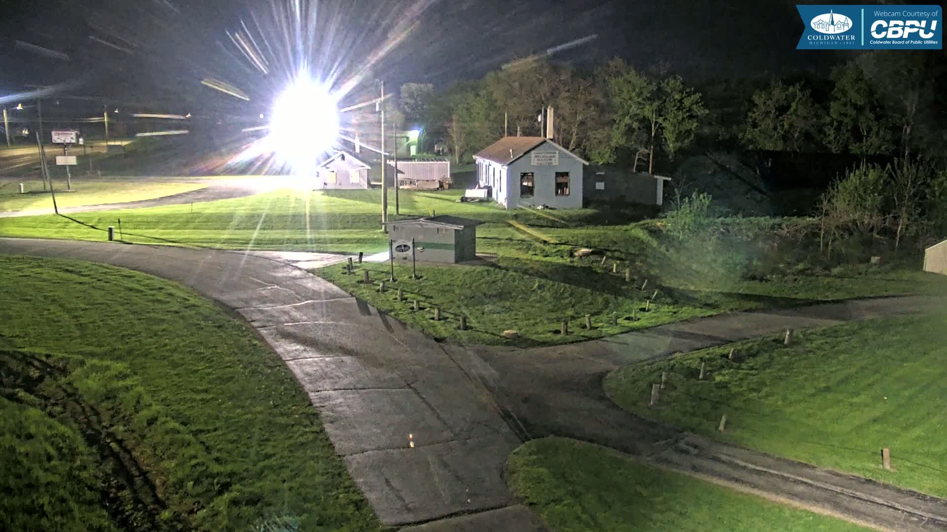 A nighttime view of a paved roadway curving through a grassy area with several small buildings and bright lights illuminating the scene.