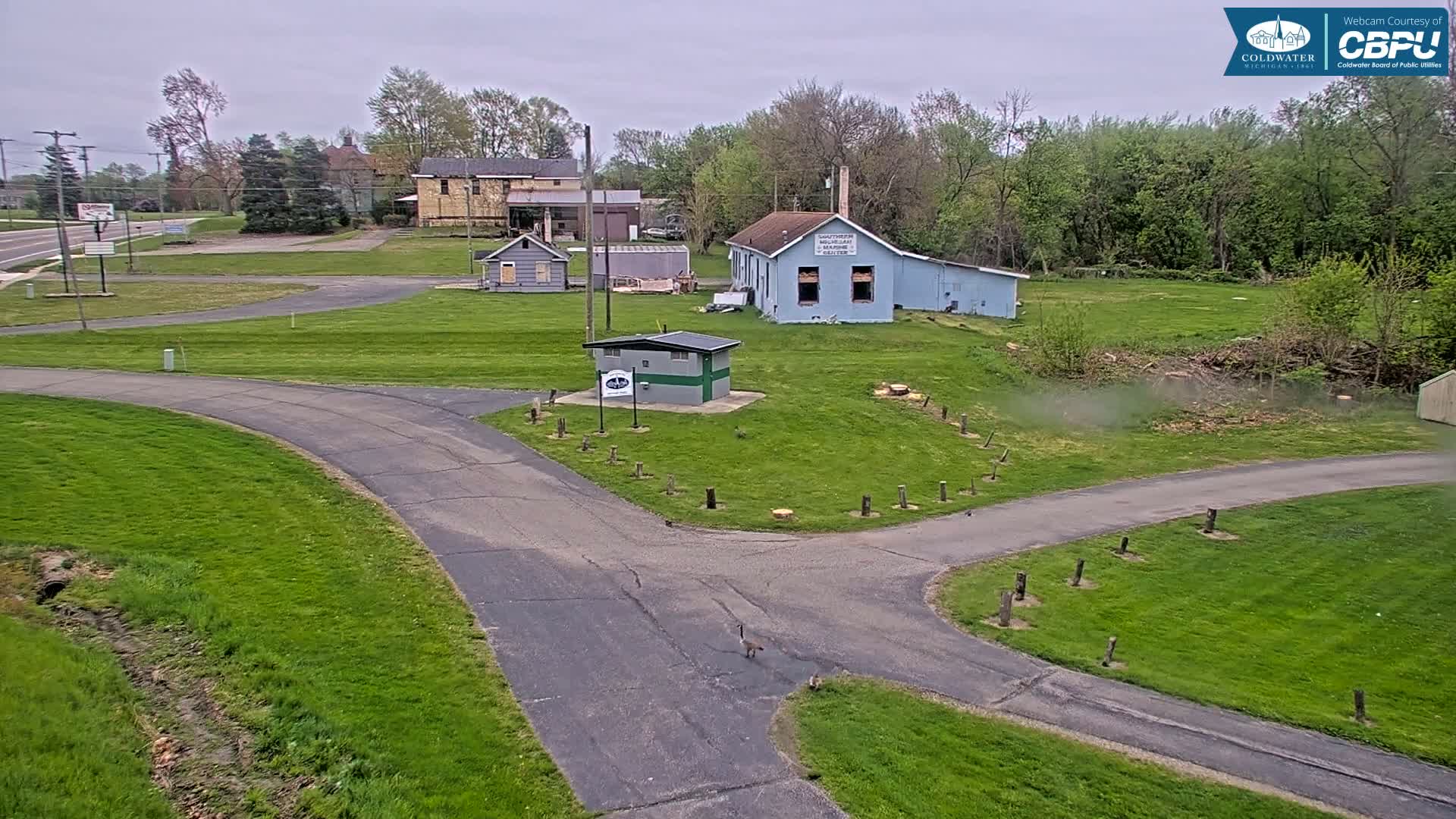 A paved roadway curves through a grassy area with several small buildings and trees under an overcast sky.