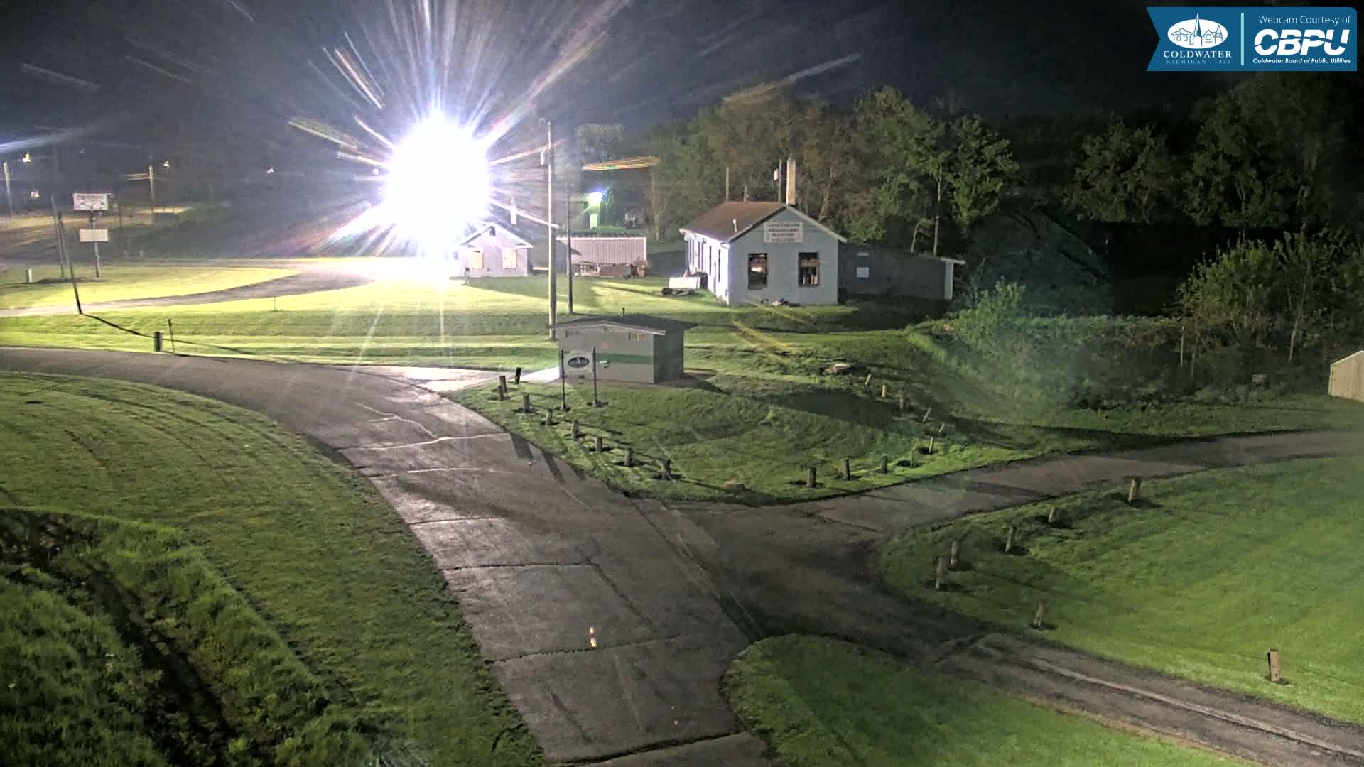 At night, under bright lights, a paved road intersects with a grassy area containing several small buildings.