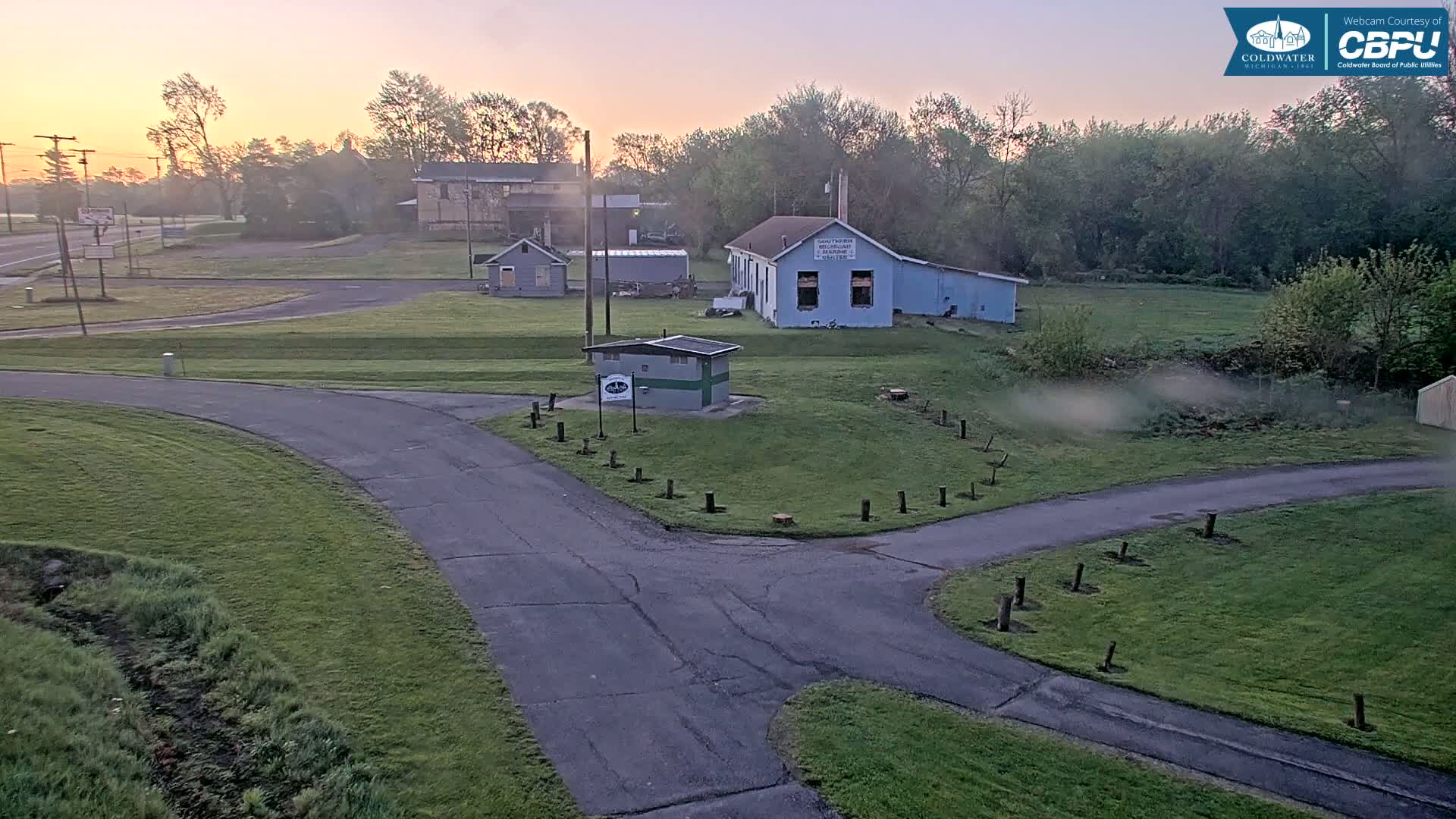 A paved road curves through a grassy area with several small buildings and trees at sunrise.