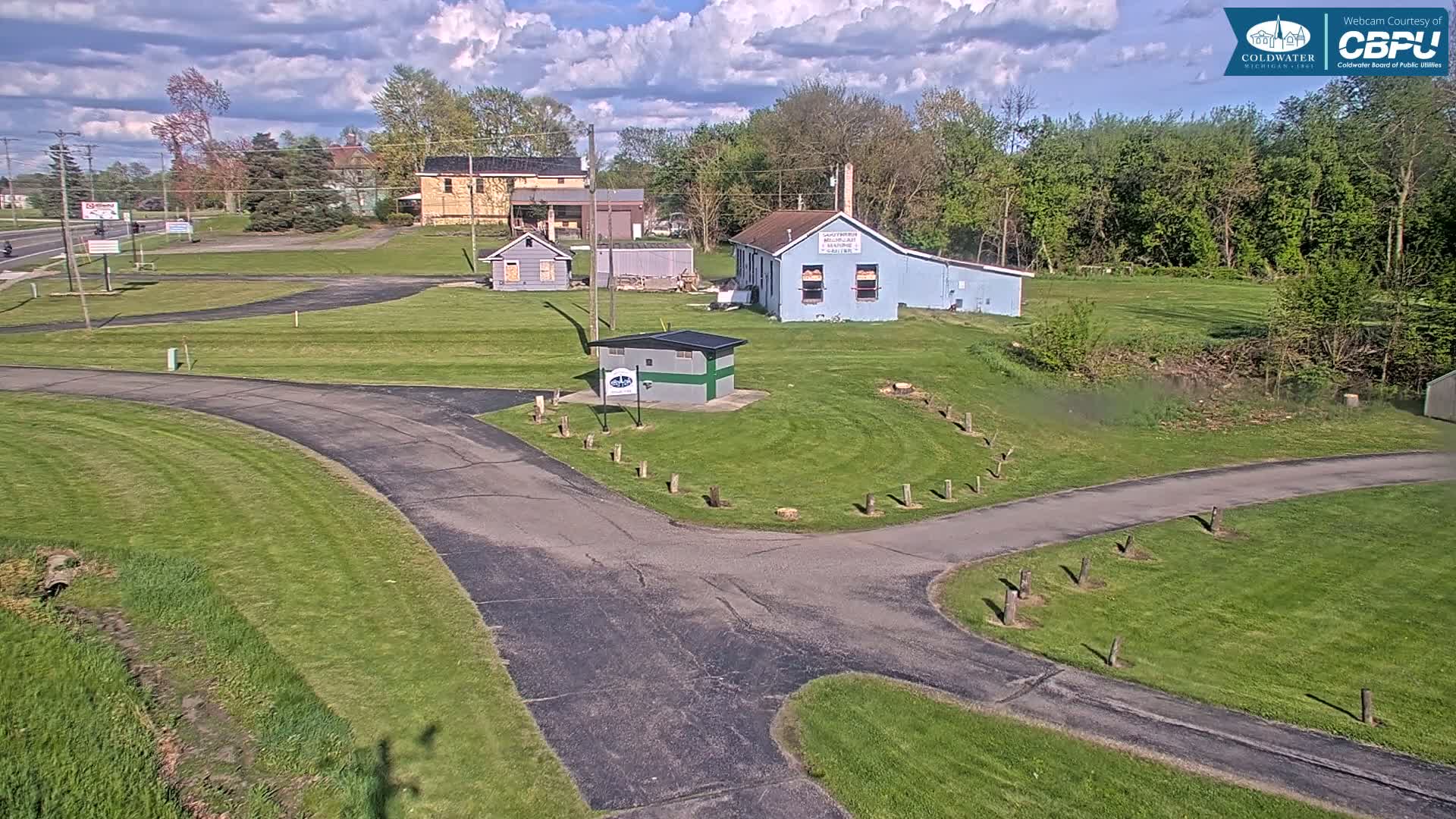 A paved area with grassy areas and several small buildings is visible under a partly cloudy sky.