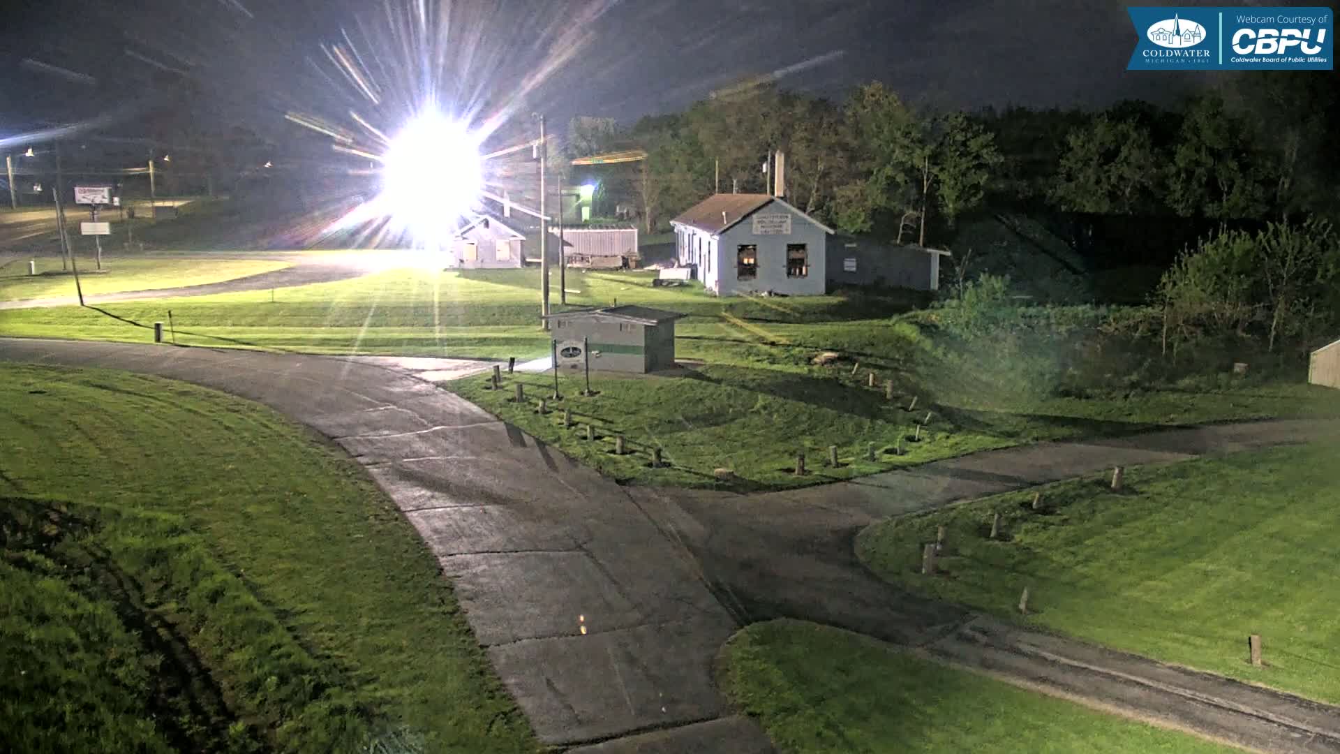 At night, under bright lights, a paved road curves through a grassy area with several small buildings and trees in the background.
