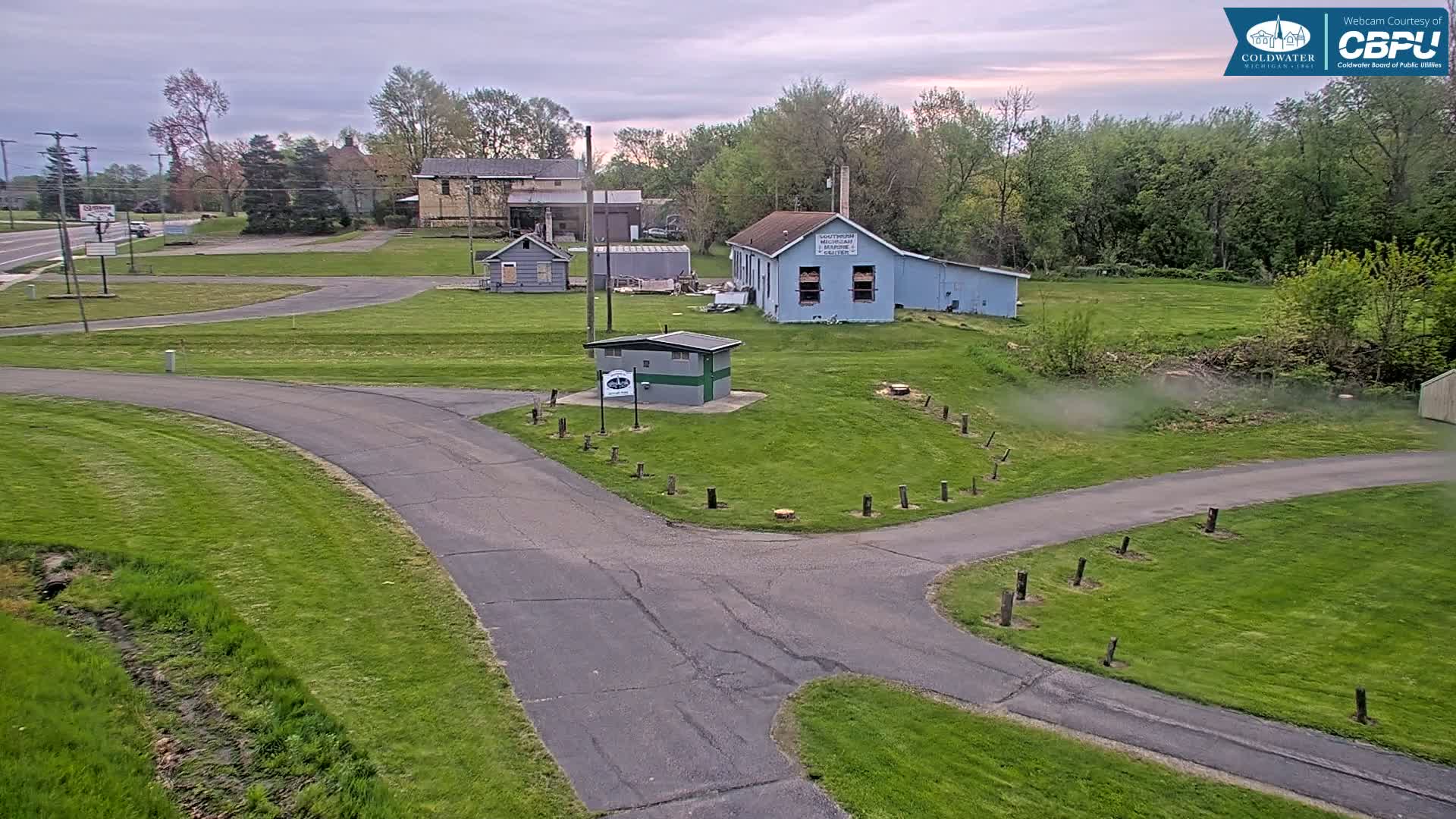 A paved roadway curves through a grassy area with several small buildings and trees under an overcast sky.