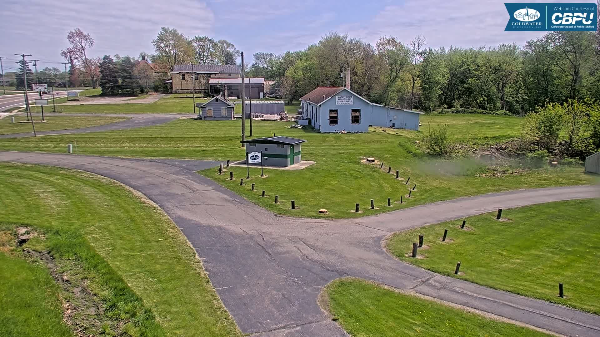 A paved pathway curves through a grassy area with several small buildings and trees under a partly cloudy sky.