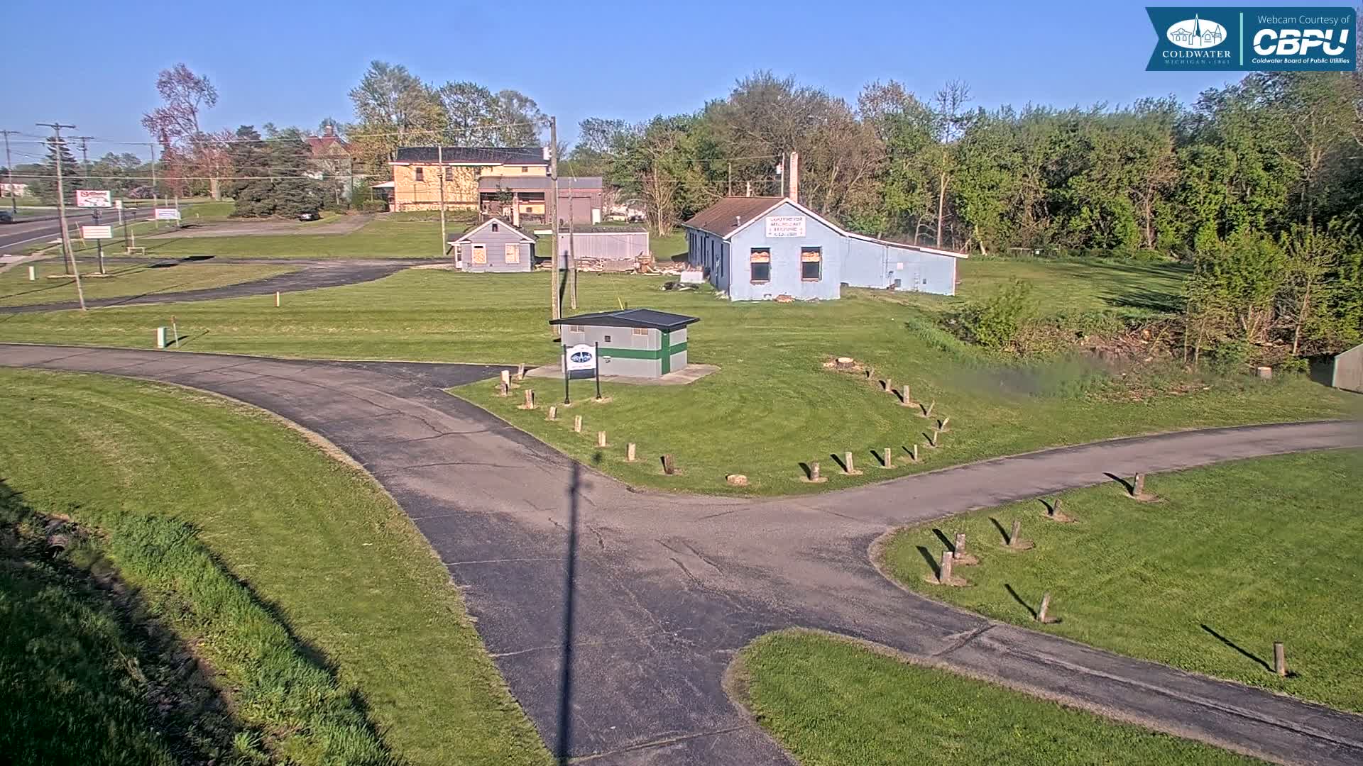A paved roadway curves through a grassy area with several small buildings and trees under a clear, sunny sky.