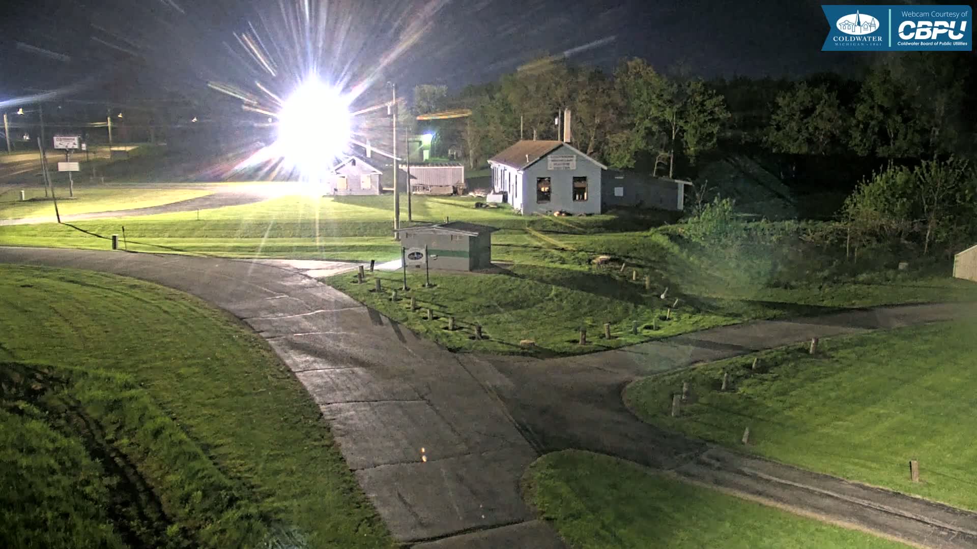 At night, under bright lights, paved roads curve through a grassy area with several small buildings, surrounded by trees.