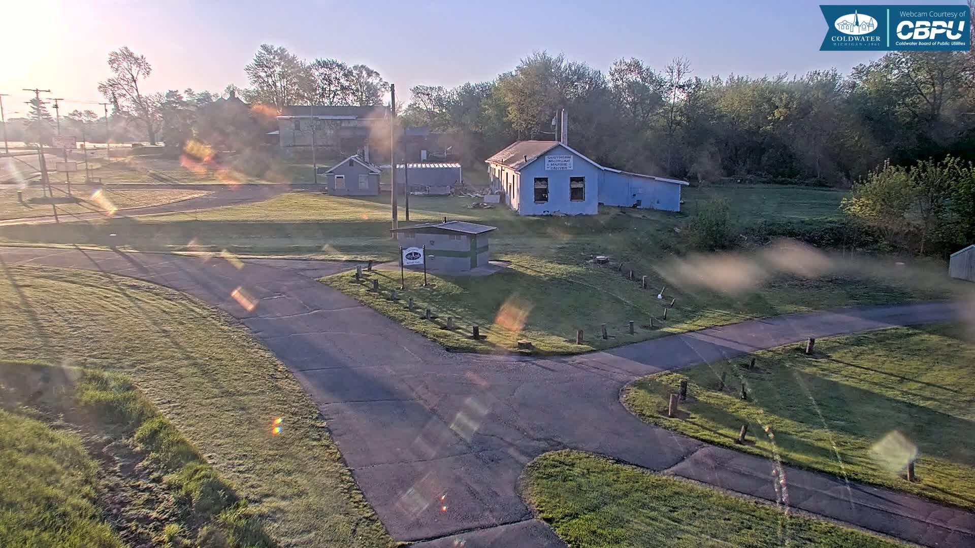 A sunny morning reveals a grassy area with several small buildings, paved paths, and trees in the background.