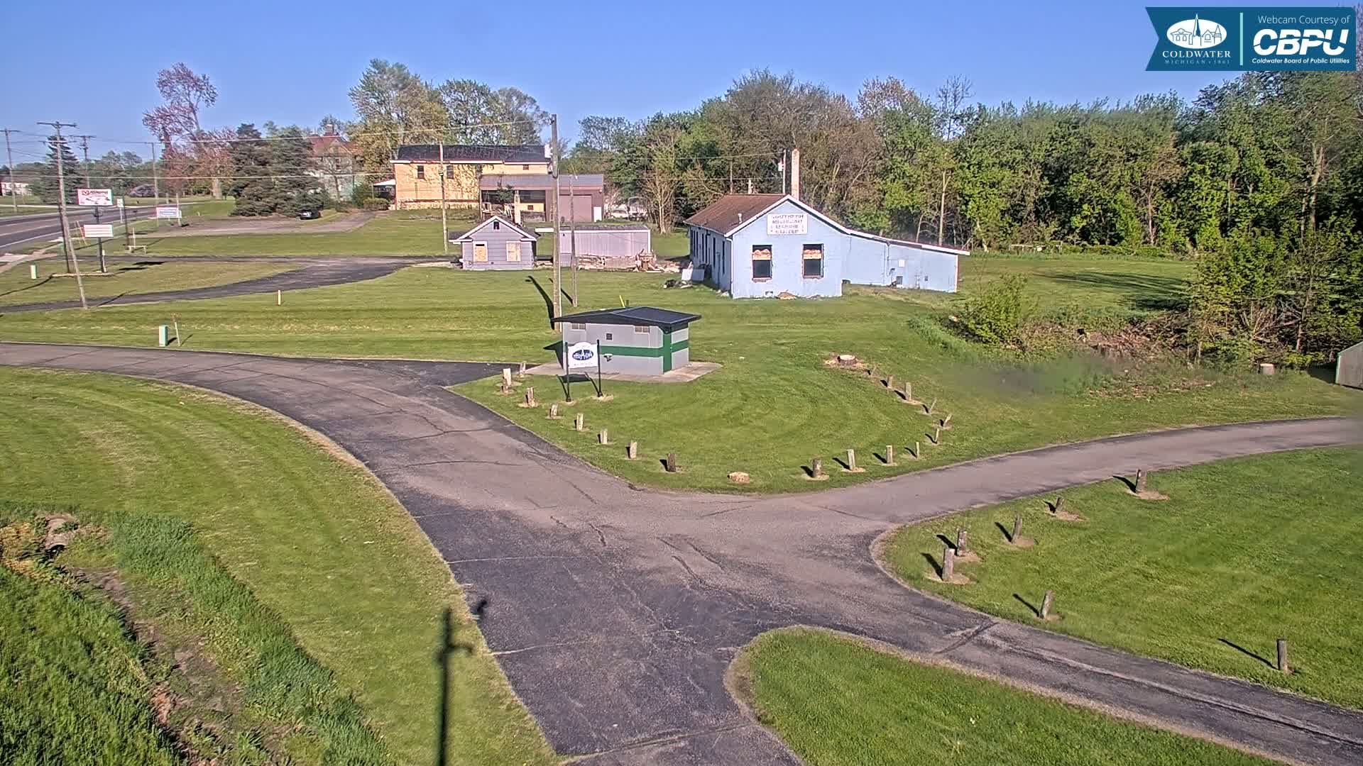 A paved roadway curves through a grassy area with several small buildings and trees under a sunny sky.