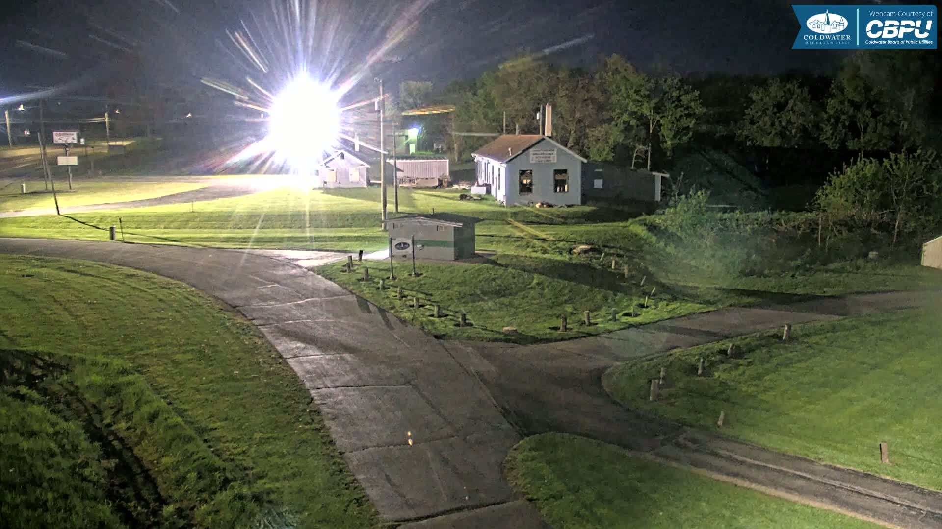 At night, a brightly lit area shows a paved road intersecting with another road, surrounded by grassy areas and a few small buildings, under a clear sky.