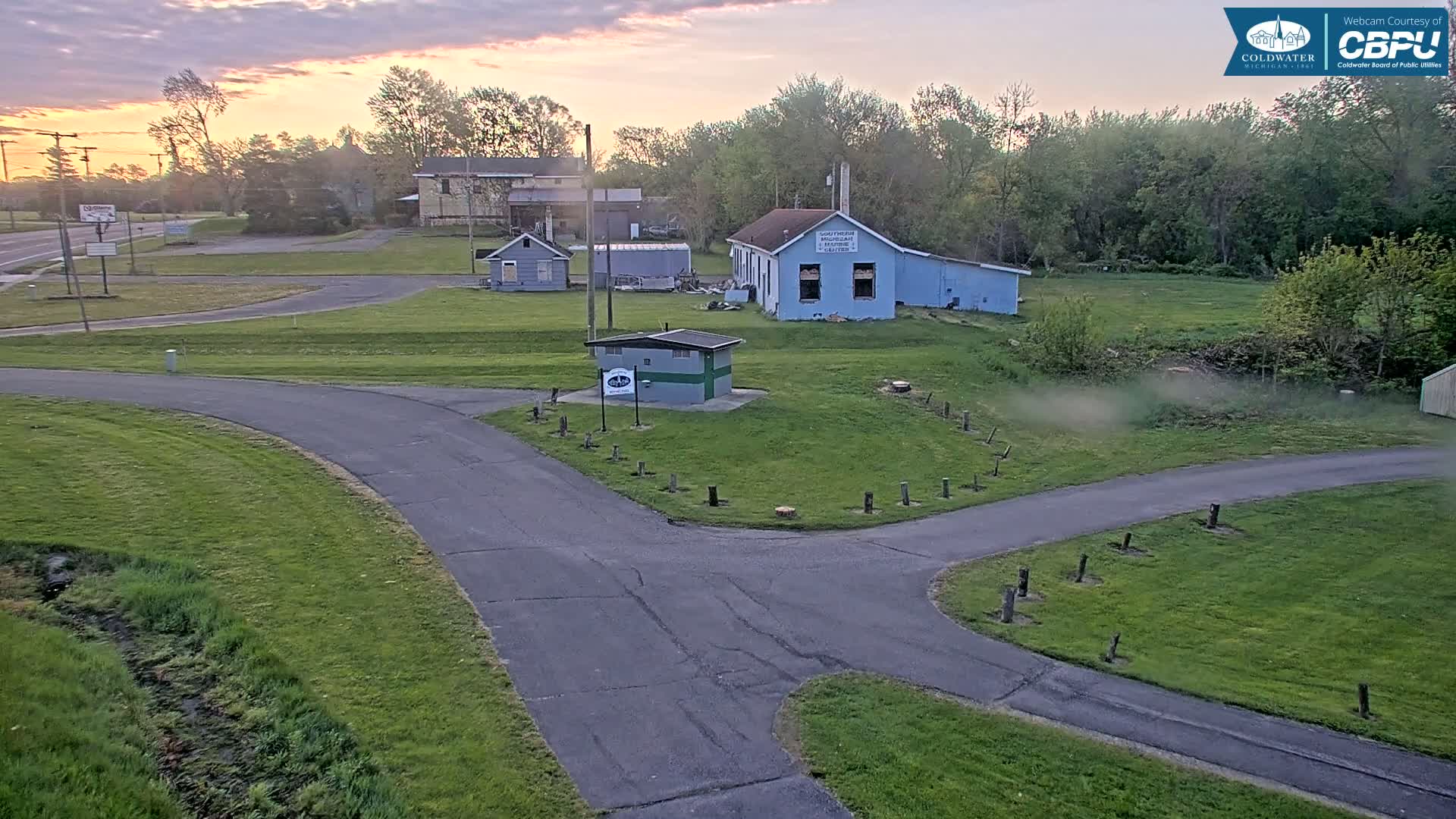 A paved road curves through a grassy area with several small buildings, under a partly cloudy sunrise sky.