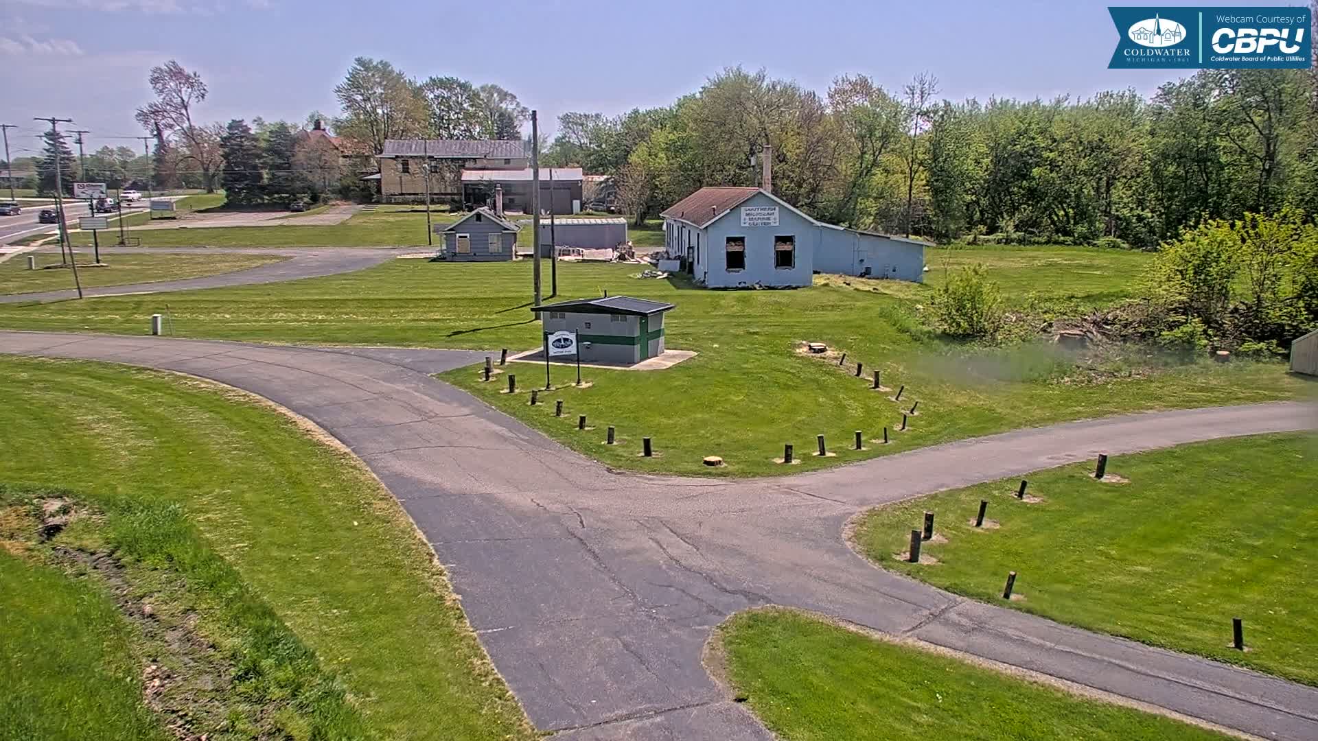 A paved path curves through a grassy area with several small buildings and trees under a sunny sky.