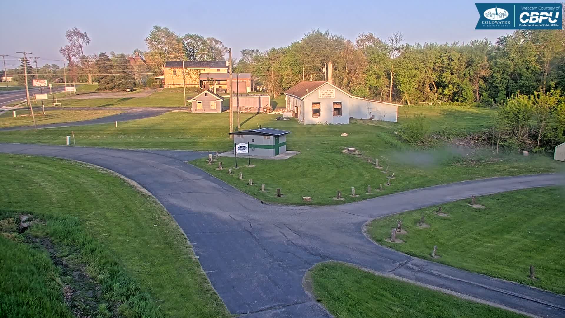 A paved road curves through a grassy area with several small buildings and trees under a clear, sunrise sky.